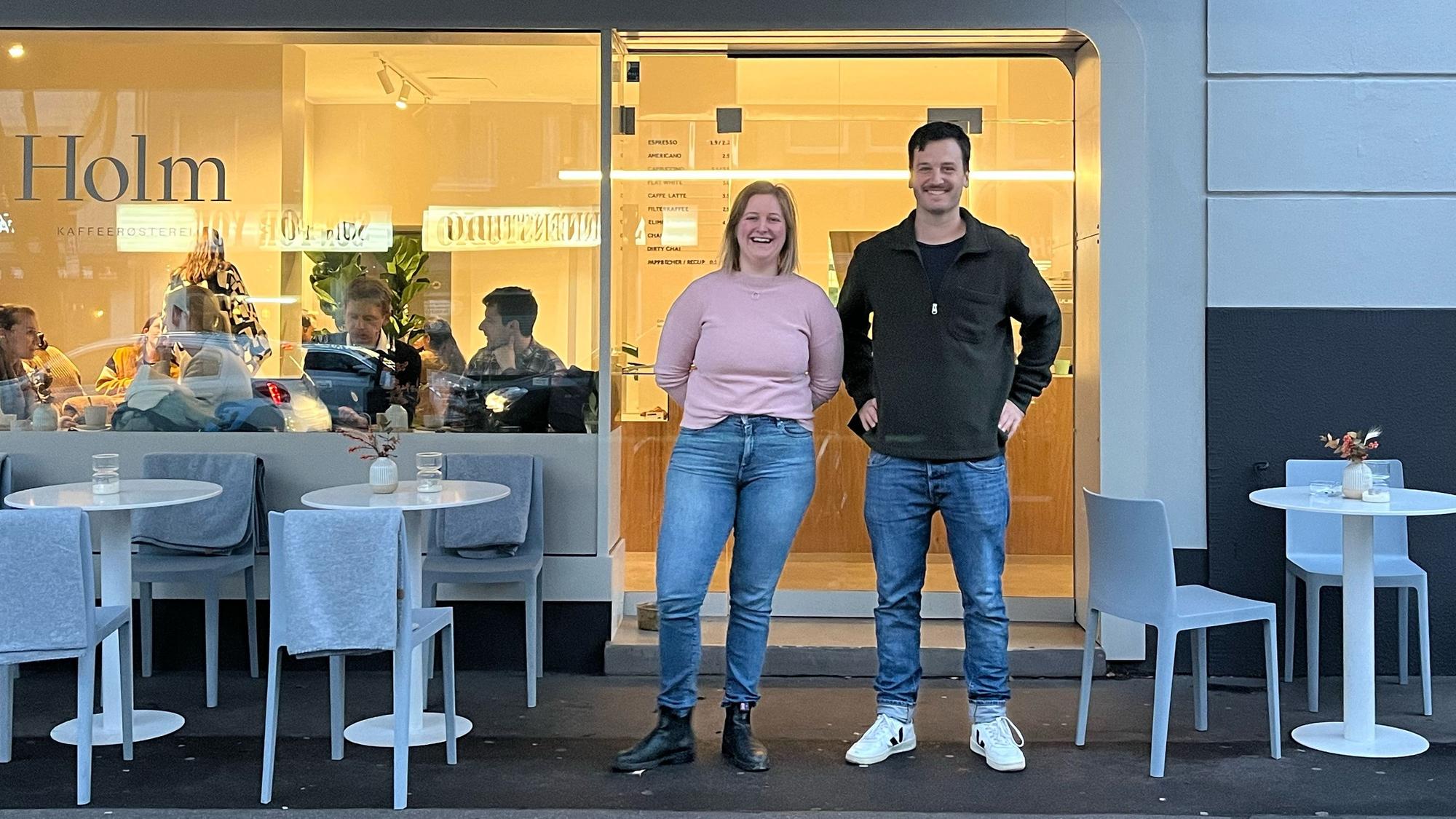 Eine Frau und ein Mann stehen vor der Glasfront eines Cafés. Links und rechts von ihnen stehen weiße Tische mit Stühlen.