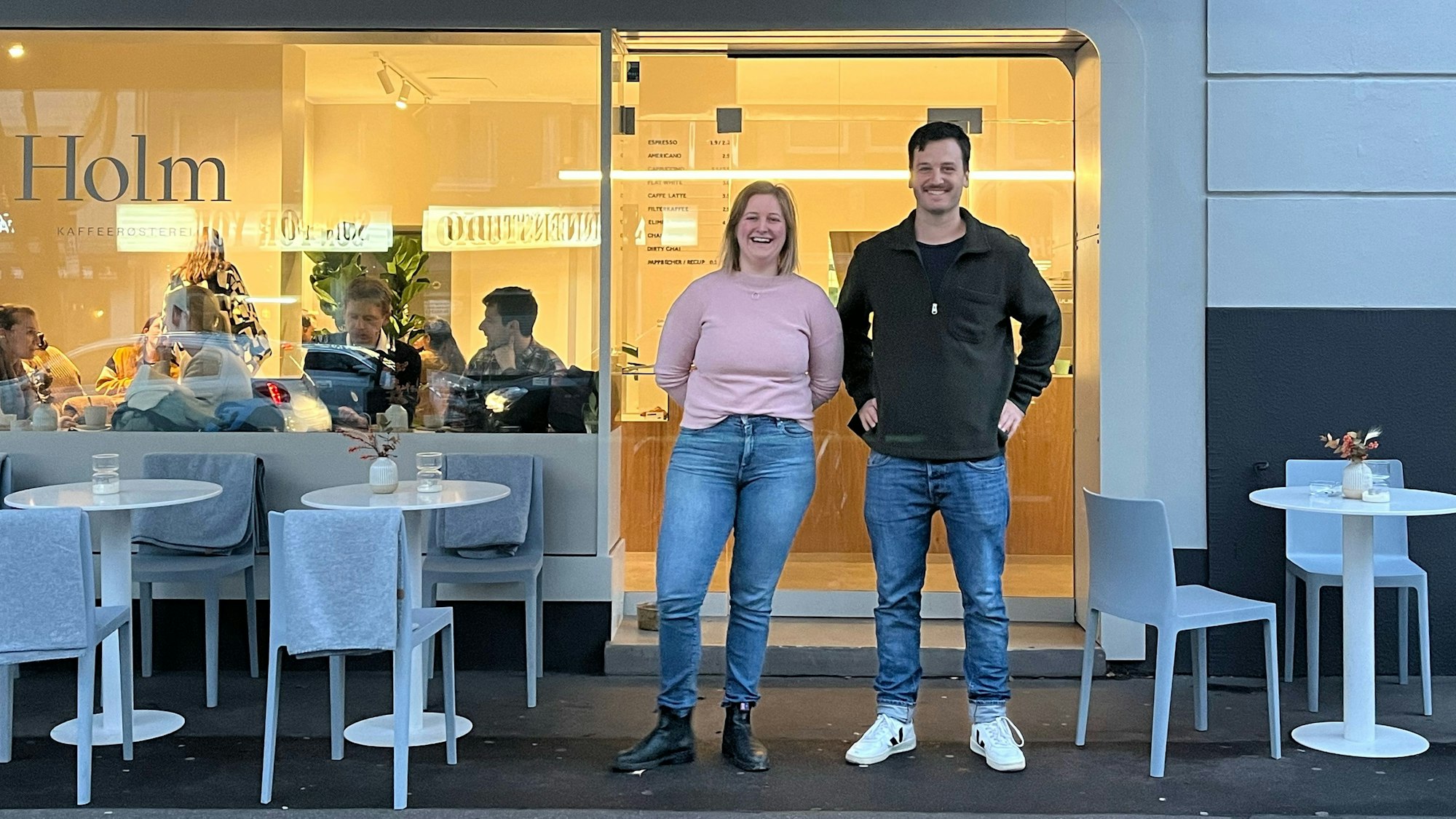 Eine Frau und ein Mann stehen vor der Glasfront eines Cafés. Links und rechts von ihnen stehen weiße Tische mit Stühlen.
