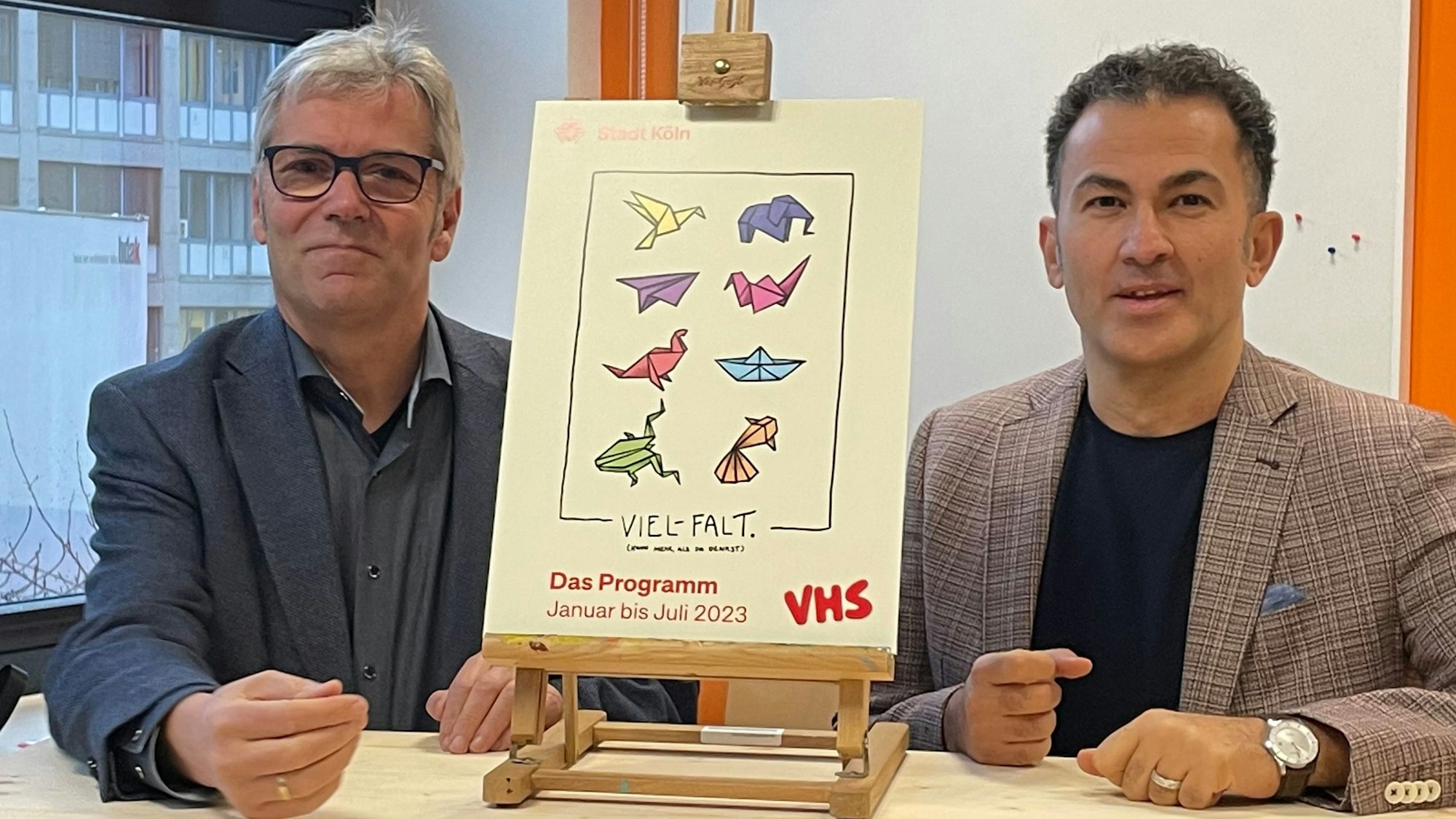 Jakob Schüller (l.) und Fatih Cevikkollu sitzen an einem Tisch. Zwischen ihnen steht ein kleines Plakat mit dem Halbjahresprogramm der VHS.