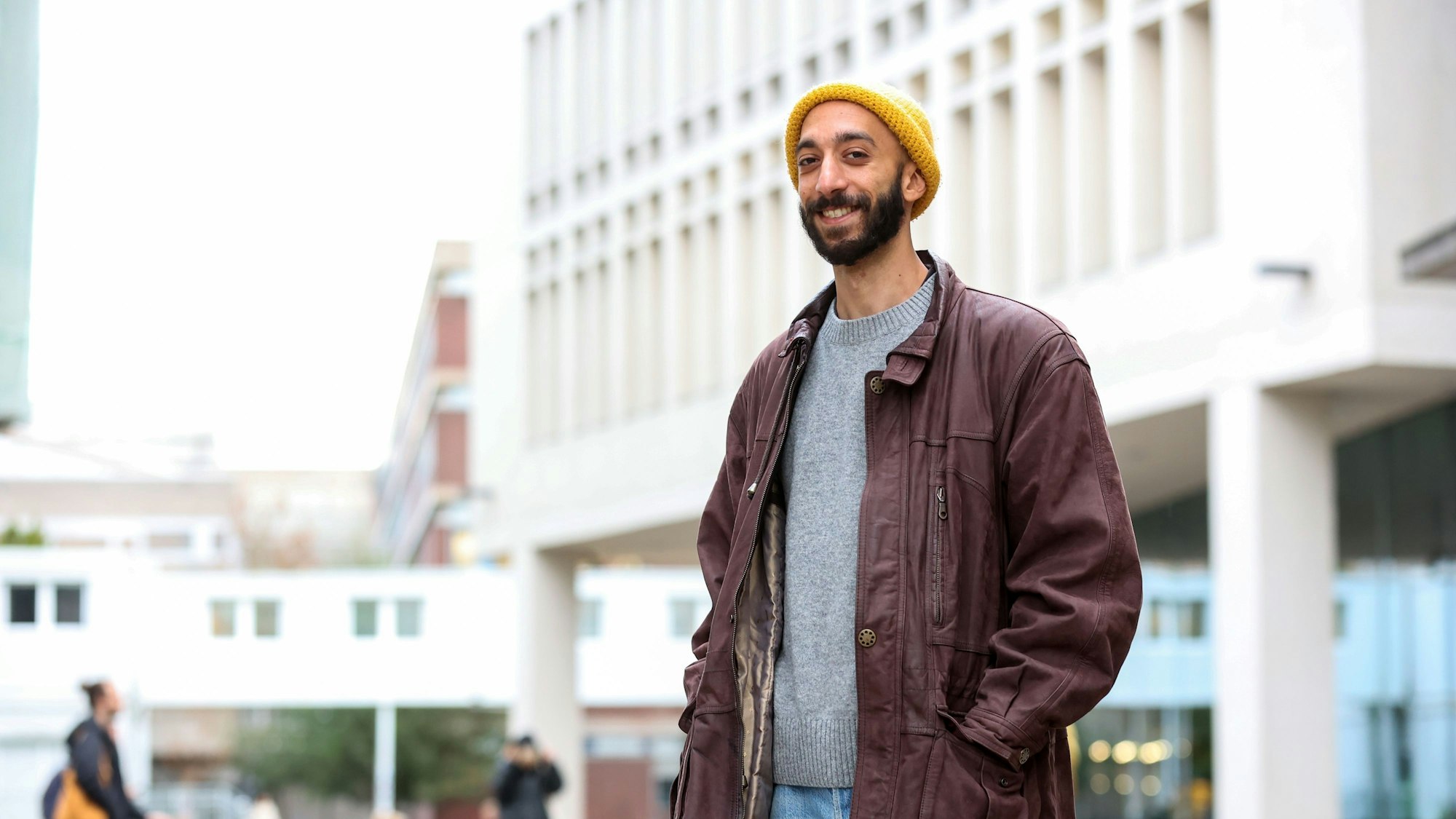 Youssef Mahfouz lehnt in einem Mantel in dunkel-lila und mit gelber Strickmütze vor der Universität zu Köln.