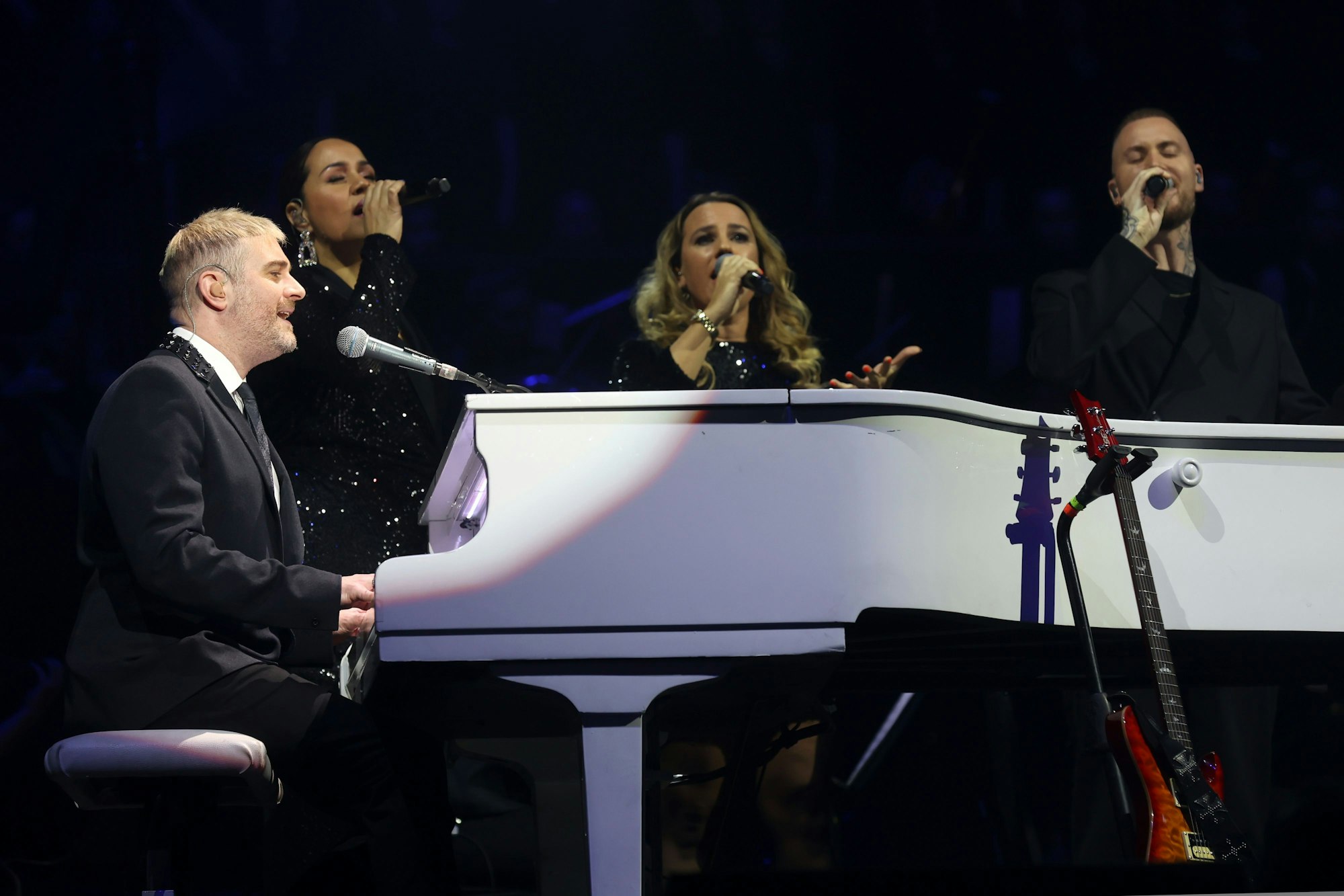 John Miles Jr. sitzt vor einem weißen Klavier und singt in ein Mikrofon.