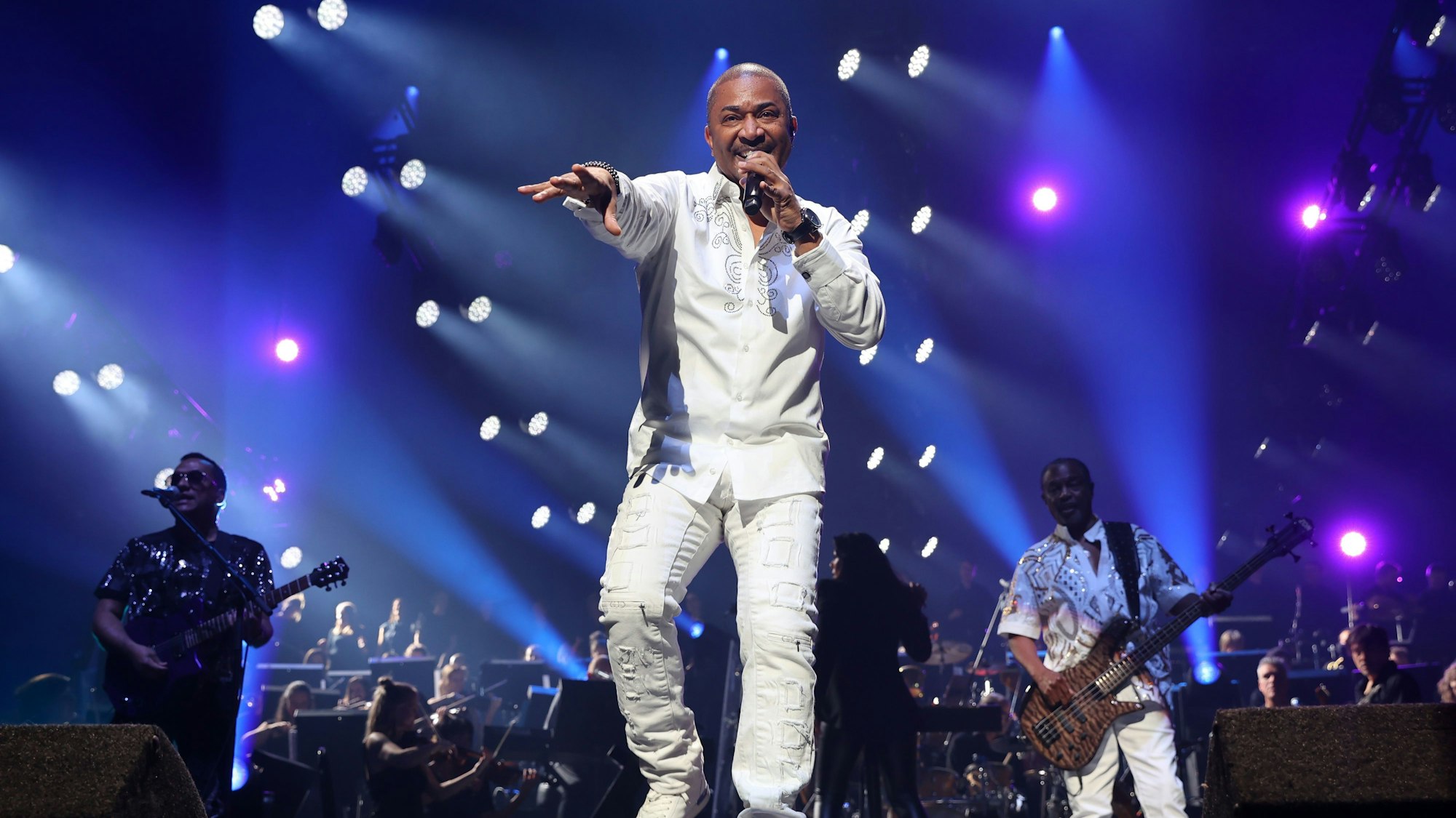 Sänger Shawn McQuiller in weißer Hose und weißem Hemd singt ins Mikrofon, das er mit einer Hand festhält. Mit der anderen zeigt er nach vorne.