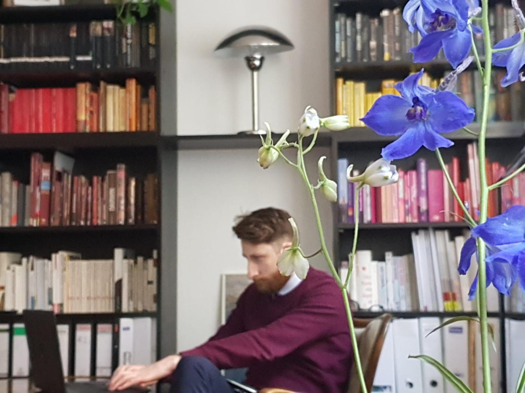 James Hargreaves sitzt in einem roten Pullover am Laptop vor einem Regal mit Büchern.
