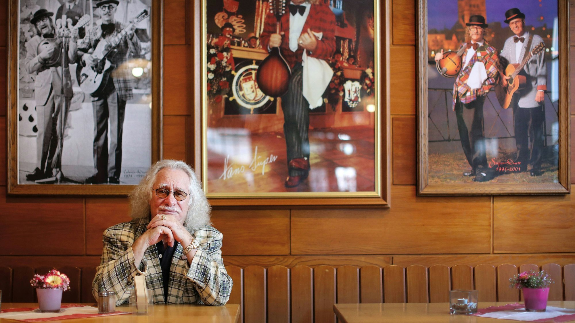 Hans Süper sitzt an einem Kneipentisch, hinter ihm Bilder von ihm bei Bühnen-Auftritten.