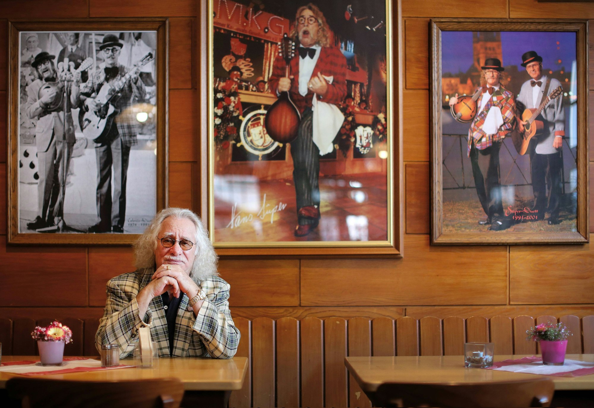 Hans Süper sitzt an einem Kneipentisch, hinter ihm Bilder von ihm bei Bühnen-Auftritten.