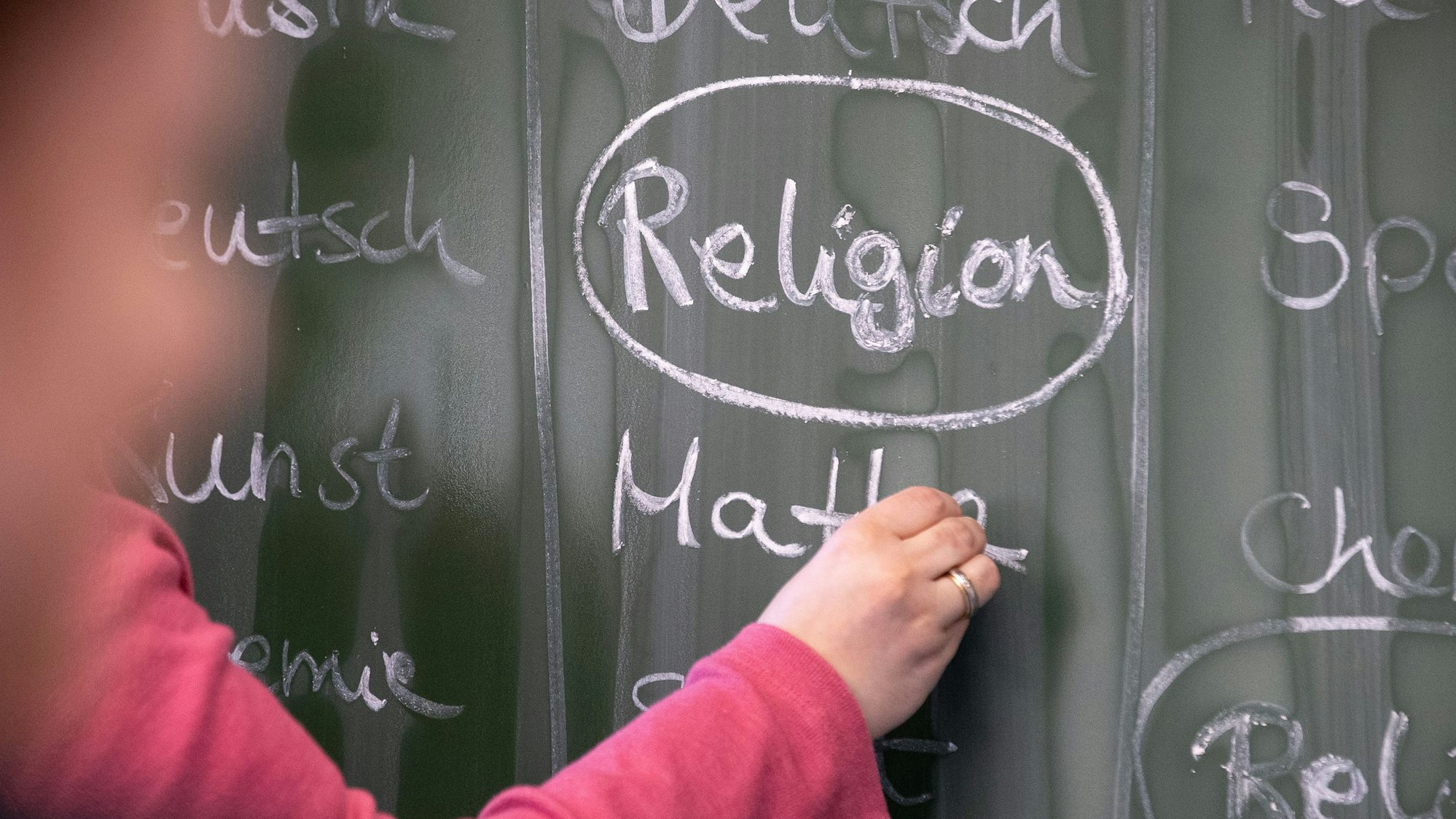 Eine Lehrerin schreibt mit Kreide einen Stundenplan mit Religionsunterricht an eine Tafel.
