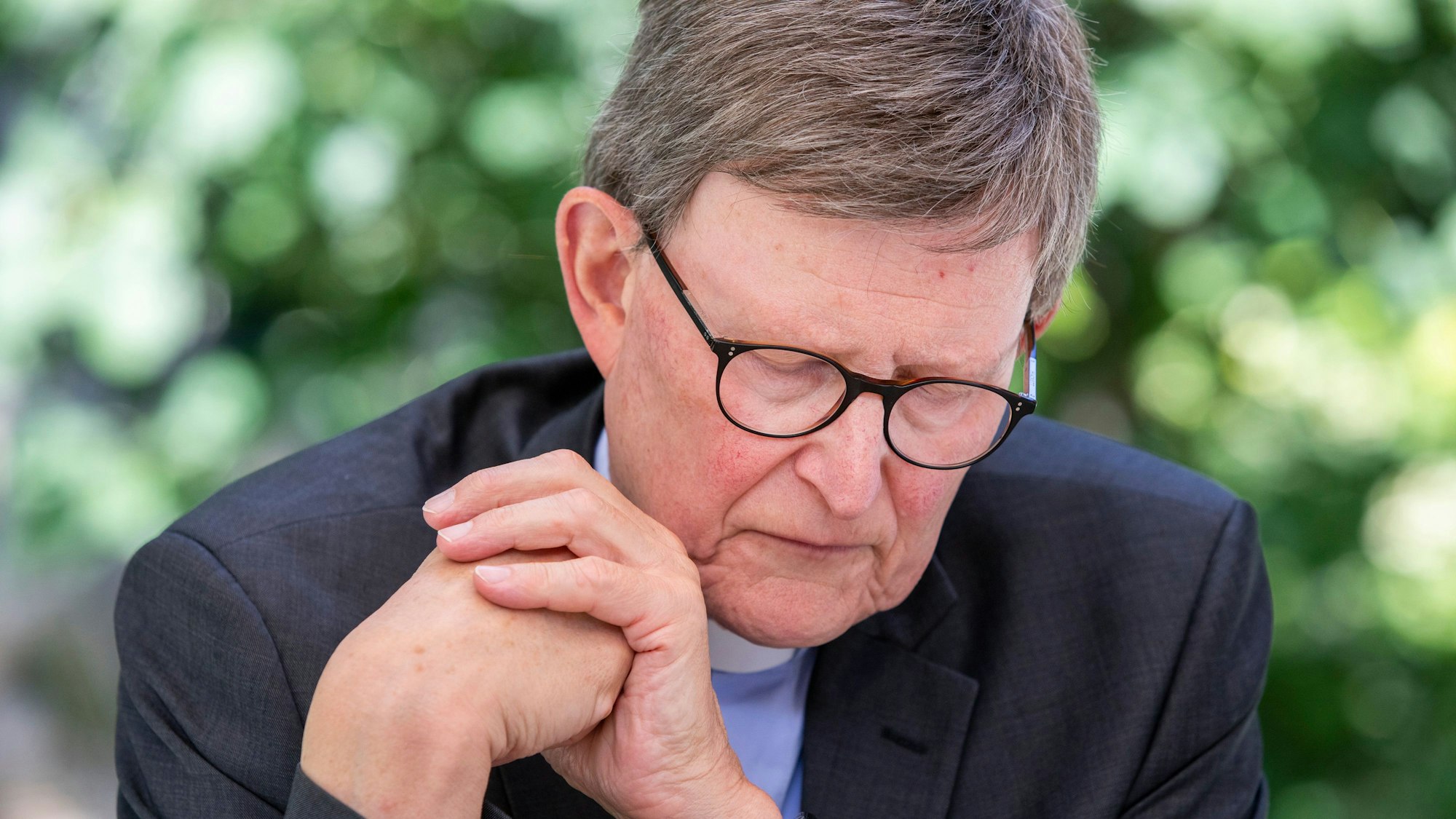Der Kölner Erzbischof, Kardinal Rainer Woelki, sitzt mit gesenktem Blick und ineinander geschlagenen Händen im Garten des erzbischöflichen Hauses.