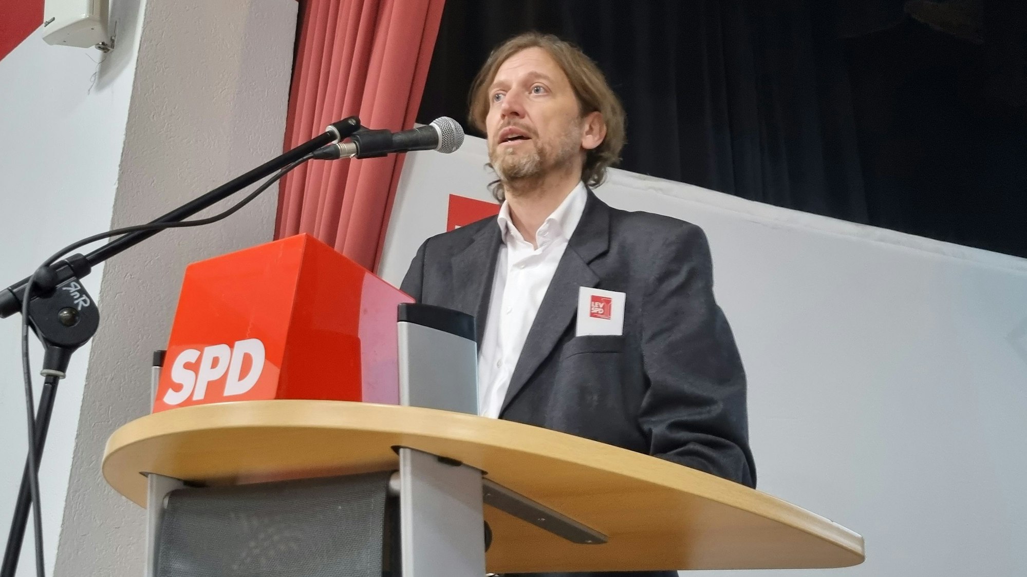 Alexander Finke, ein großer Mann mit mittellangen Haaren und Bart, steht an einem Sprechpult, vor ihm ist das Logo der SPD zu sehen.