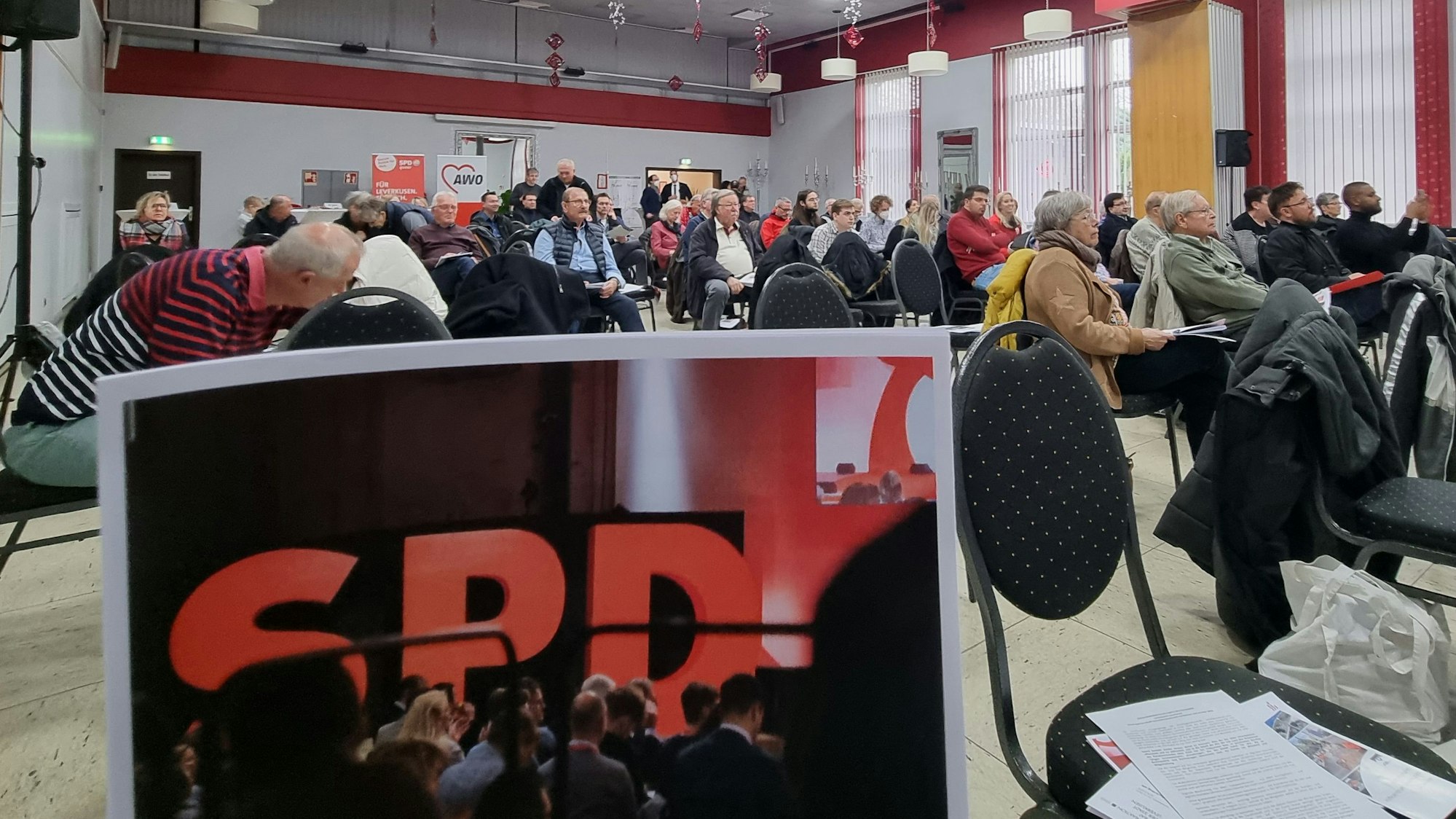 Mitglieder der SPD Leverkusen sitzen in einem großen Saal und schauen auf eine Bühne. Im Vordergrund ist ein Foto zu sehen, auf dem der Schriftzug SPD steht.