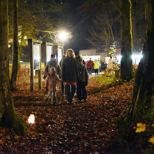 Eine Familie wandert durch einen dunkeln Wald, zwischen den Bäumen sind Teelichter aufgestellt.