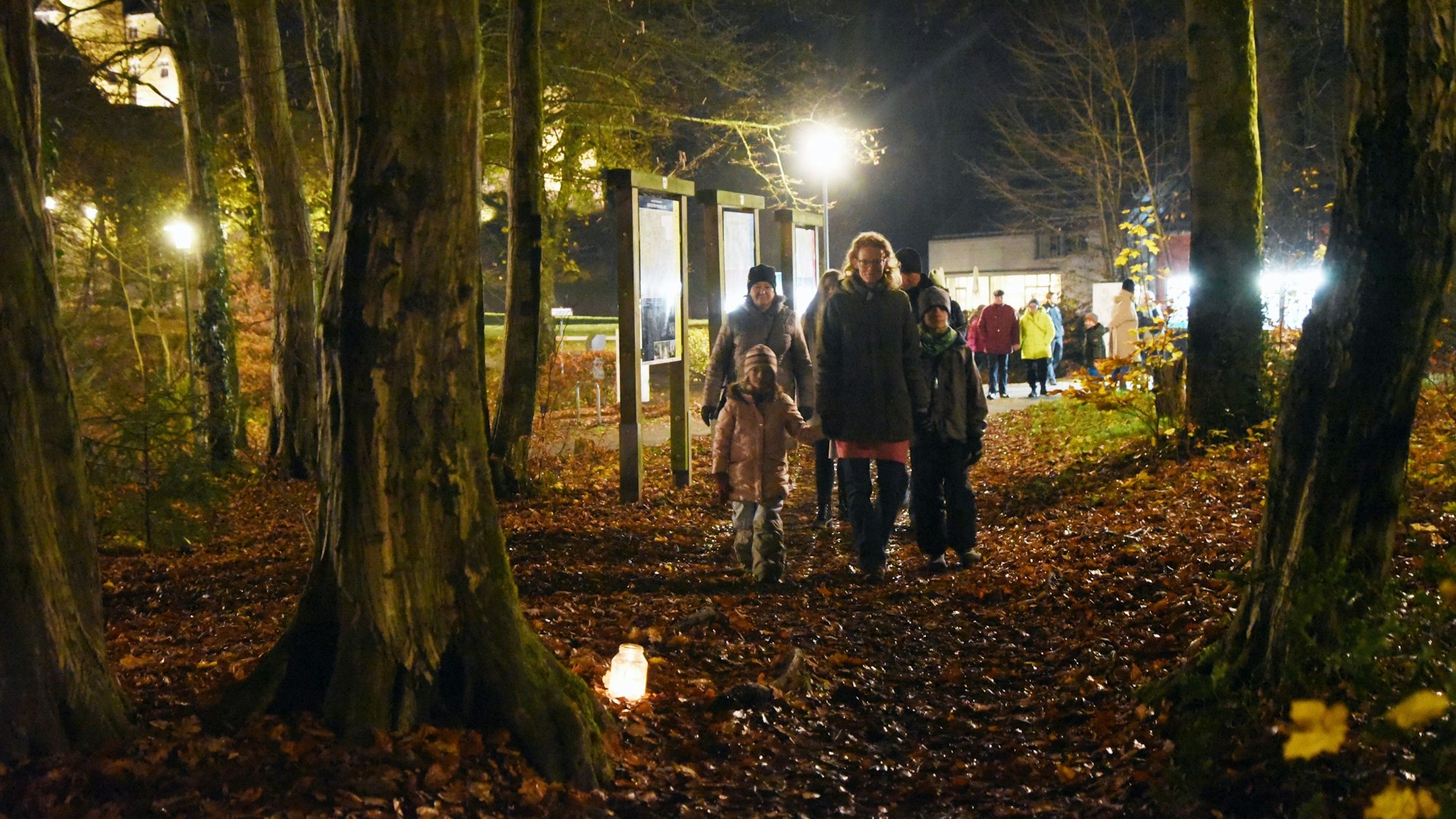 Eine Familie wandert durch einen dunkeln Wald, zwischen den Bäumen sind Teelichter aufgestellt.