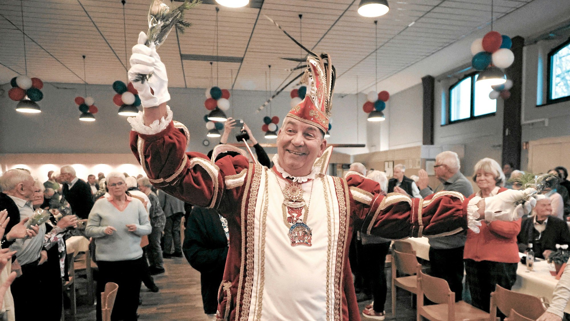Hitdorfs Prinz Karneval Volker I. läuft in vollem Ornat lachend durch den Gang, links und rechts von ihm befinden sich Tische und Stühle und vielen Seniorinnen und Senioren.