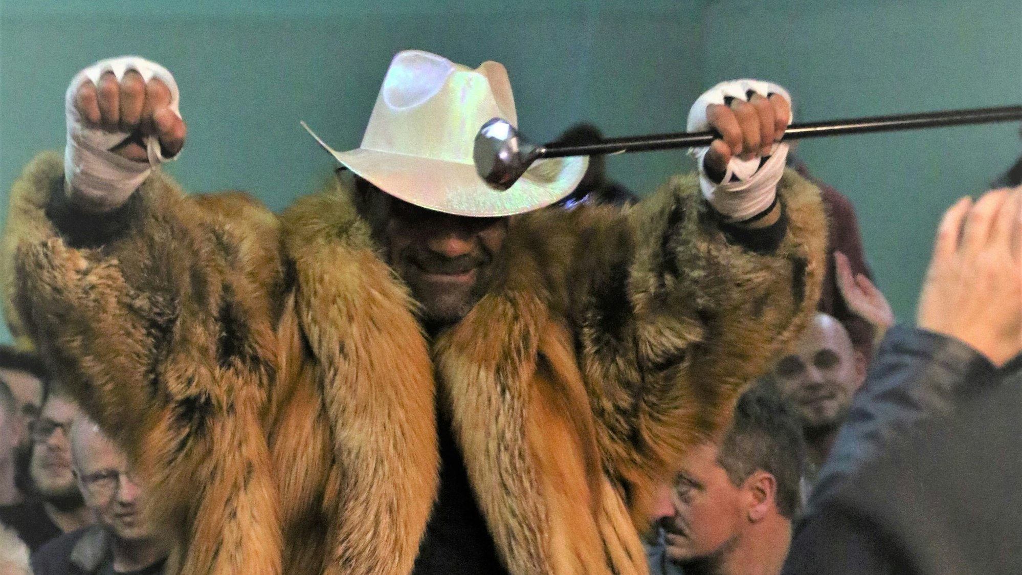 Remo Arns geht mit Cowboyhut und Pelzmantel mit erhobenen Armen durch die Menge bei der Mechernicher Fight Night. In einer Hand hält er einen Gehstock.