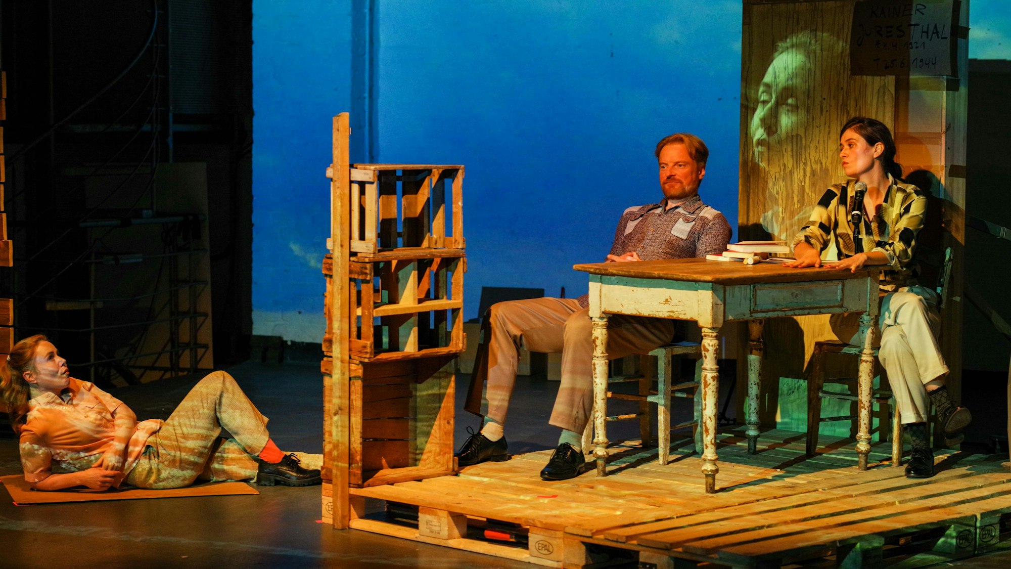 Szene aus „Annette, Ein Heldinnenepos“ im Freien Werkstatt Theater. Annette liegt auf einer Matte am Boden, darüber, auf einer Konstruktion aus Palletts, sitzen ein Mann und eine Frau an einem Tisch.
