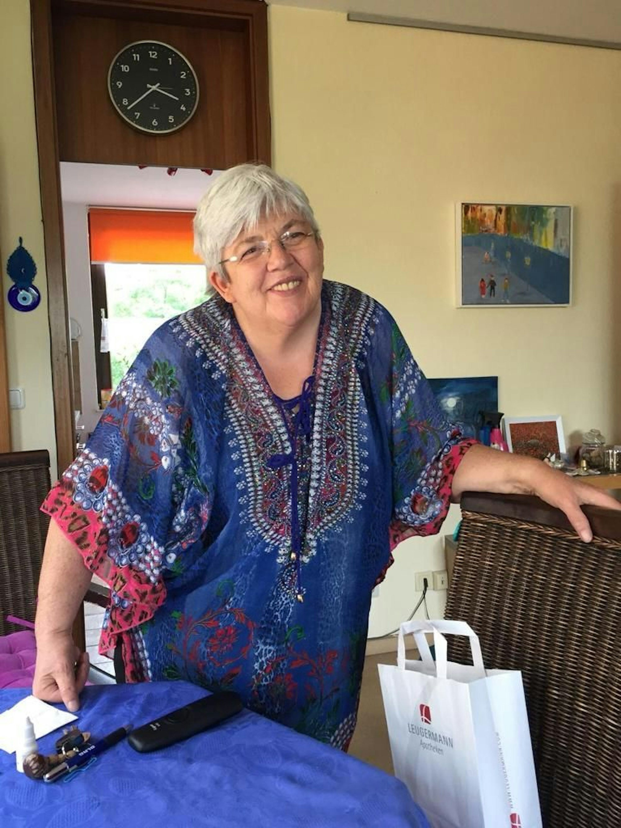 Brigitte Lersch steht mit einem einem blauen Kleid vor einem Tisch mit blauer Decke und lacht.