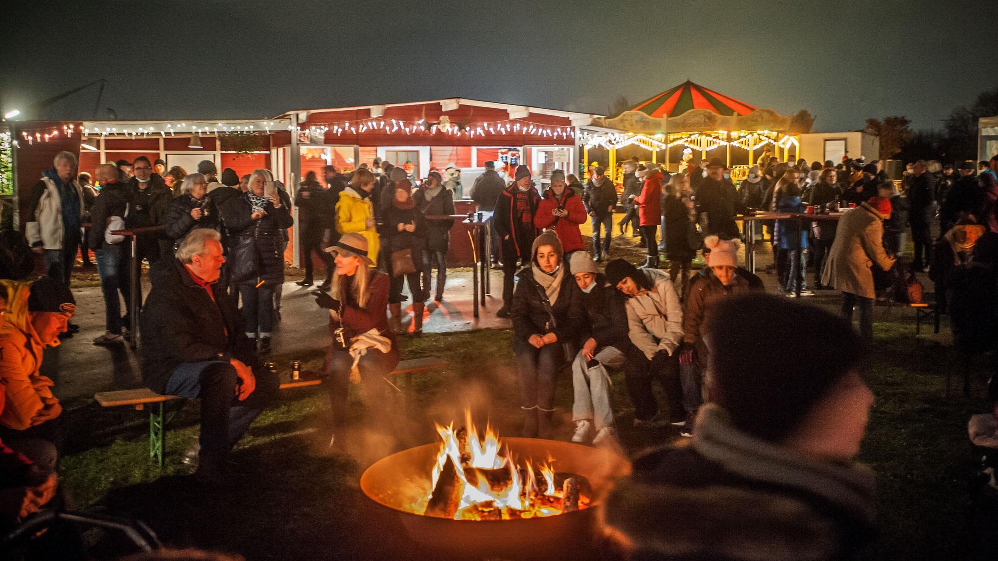 Um eine Feuerschale sitzen Besucherinnen und Besucher der Weihnachtsmarkts, im Hintergrund sind erleuchtete Buden, viele Menschen und ein Karussel zu sehen.