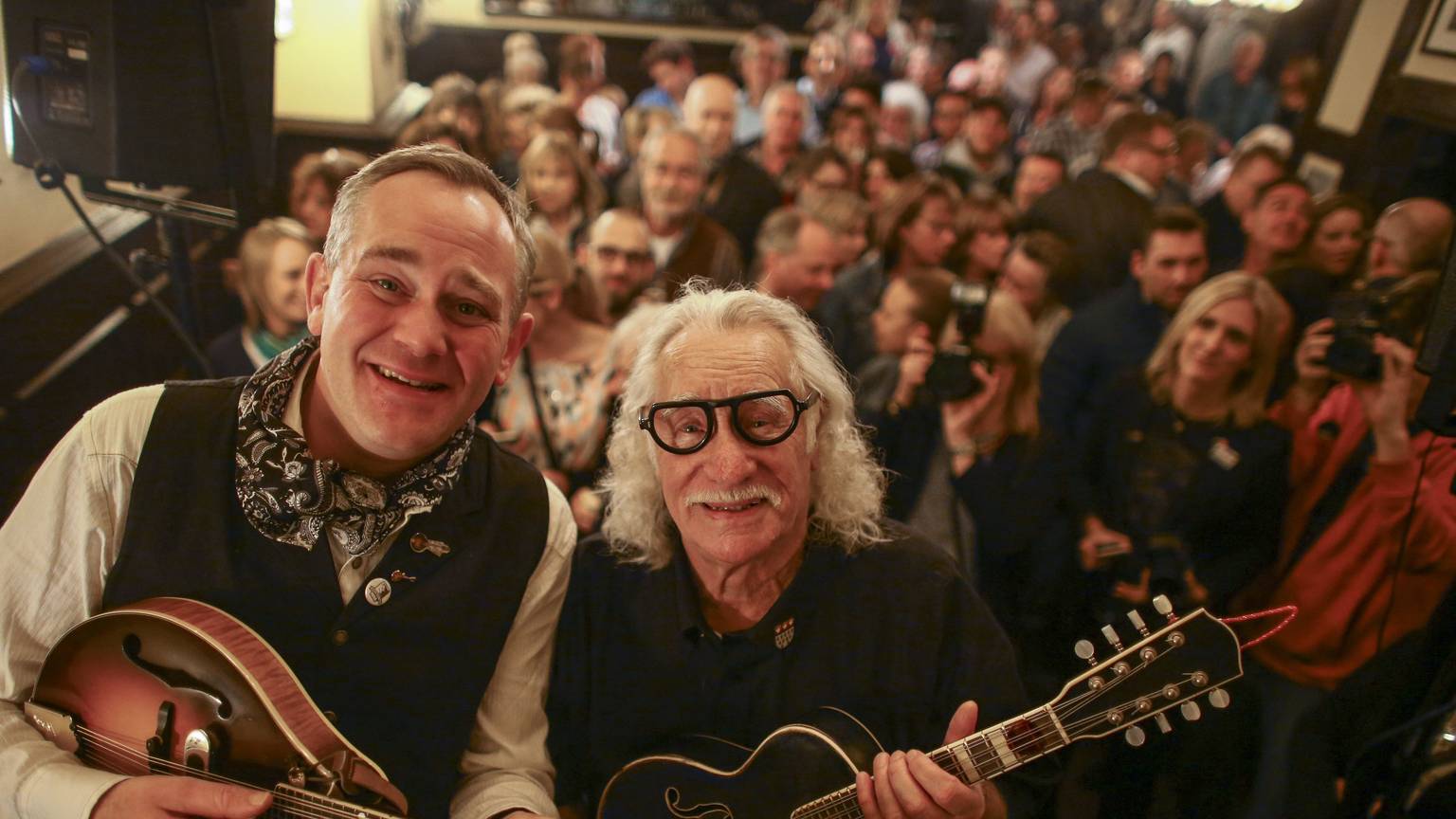 JP Weber und Hans Süper schauen mit ihren Mandolinen in den Händen in die Kamera, im Hintergrund Kneipen-Publikum.