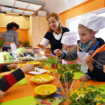 Eine Frau bereitet mit Kindergartenkindern, die Kochmützen tragen, Fruchtspieße zu.