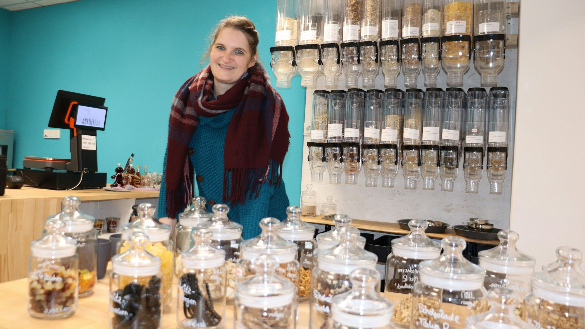 Eine Frau mit dunklem Haar steht hinter einem Verkaufstresen, auf dem Gläser mit Keksen und anderem zu sehen sind.