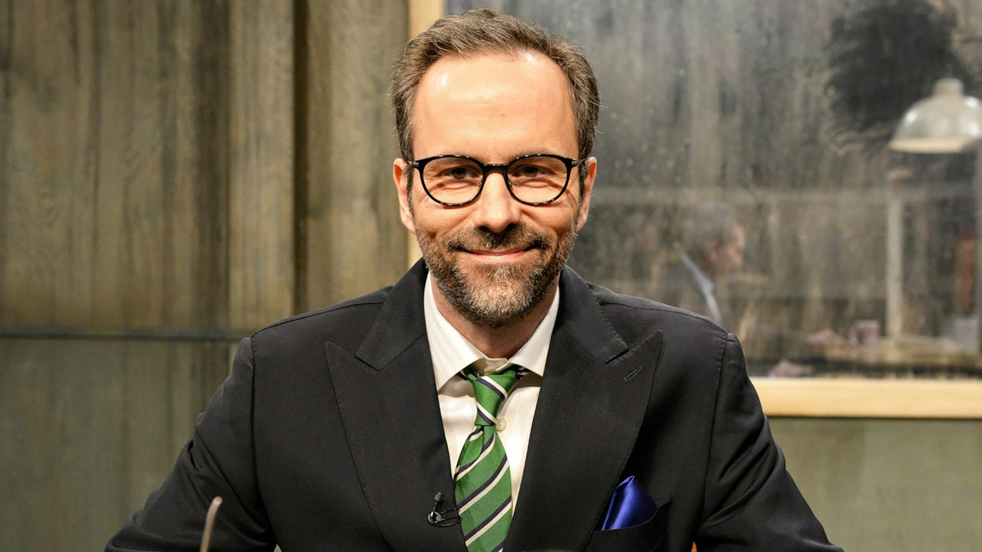 Komiker und Moderator Kurt Krömer sitzt in einem schwarzen Anzug in der Kullisse eines altmodischen Verhörraums.