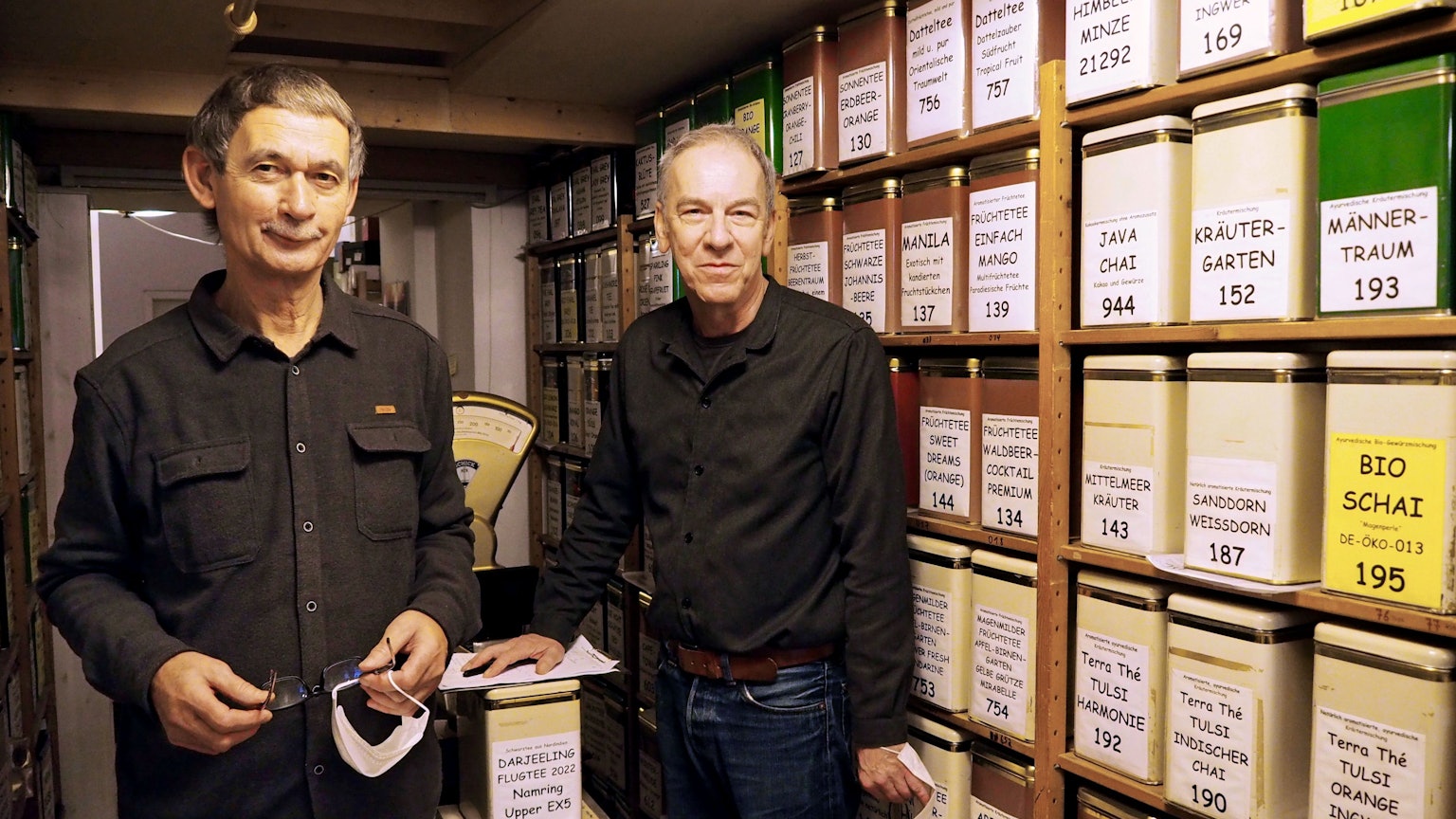 Zwei Männer um die 60 stehen neben der Wand mit den unterschiedlichen Teesorten. Sie tragen schwarze Hemden und Jeans.