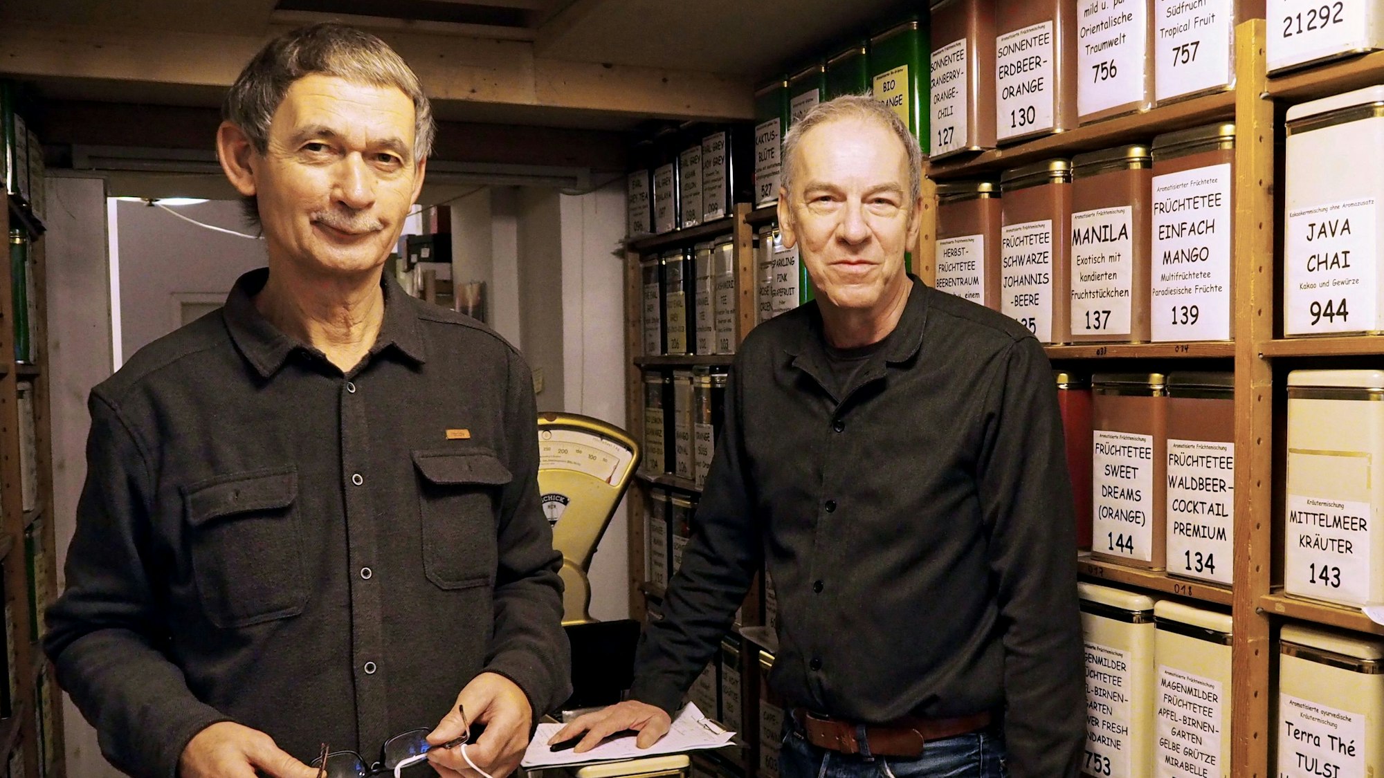 Zwei Männer um die 60 stehen neben der Wand mit den unterschiedlichen Teesorten. Sie tragen schwarze Hemden und Jeans.