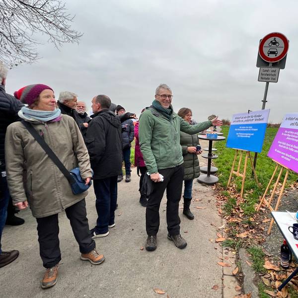 Links ist eine Gruppe von Menschen zu sehen. Einer von ihnen, Jörg Lichter, blickt in die Kamera und zeigt auf Plakat, die auf der rechten Seiten aufgetellt sind. Dort steht unter anderem „In Hitdorf wüten Bau-Wahnsinn und Flächenfraß!“