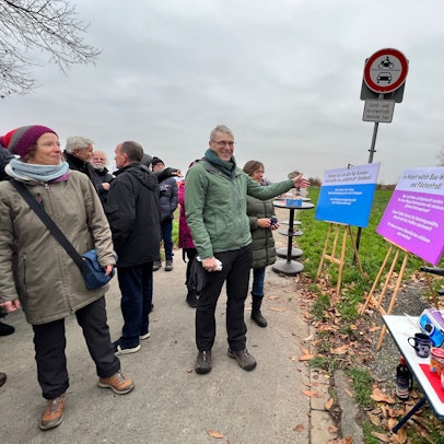 Links ist eine Gruppe von Menschen zu sehen. Einer von ihnen, Jörg Lichter, blickt in die Kamera und zeigt auf Plakat, die auf der rechten Seiten aufgetellt sind. Dort steht unter anderem „In Hitdorf wüten Bau-Wahnsinn und Flächenfraß!“
