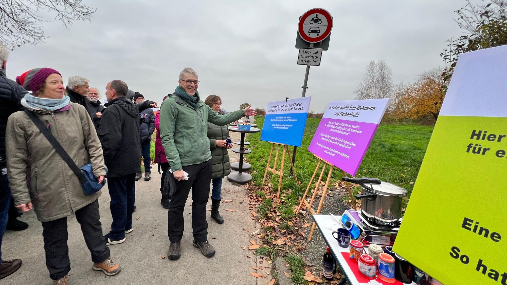 Links ist eine Gruppe von Menschen zu sehen. Einer von ihnen, Jörg Lichter, blickt in die Kamera und zeigt auf Plakat, die auf der rechten Seiten aufgetellt sind. Dort steht unter anderem „In Hitdorf wüten Bau-Wahnsinn und Flächenfraß!“