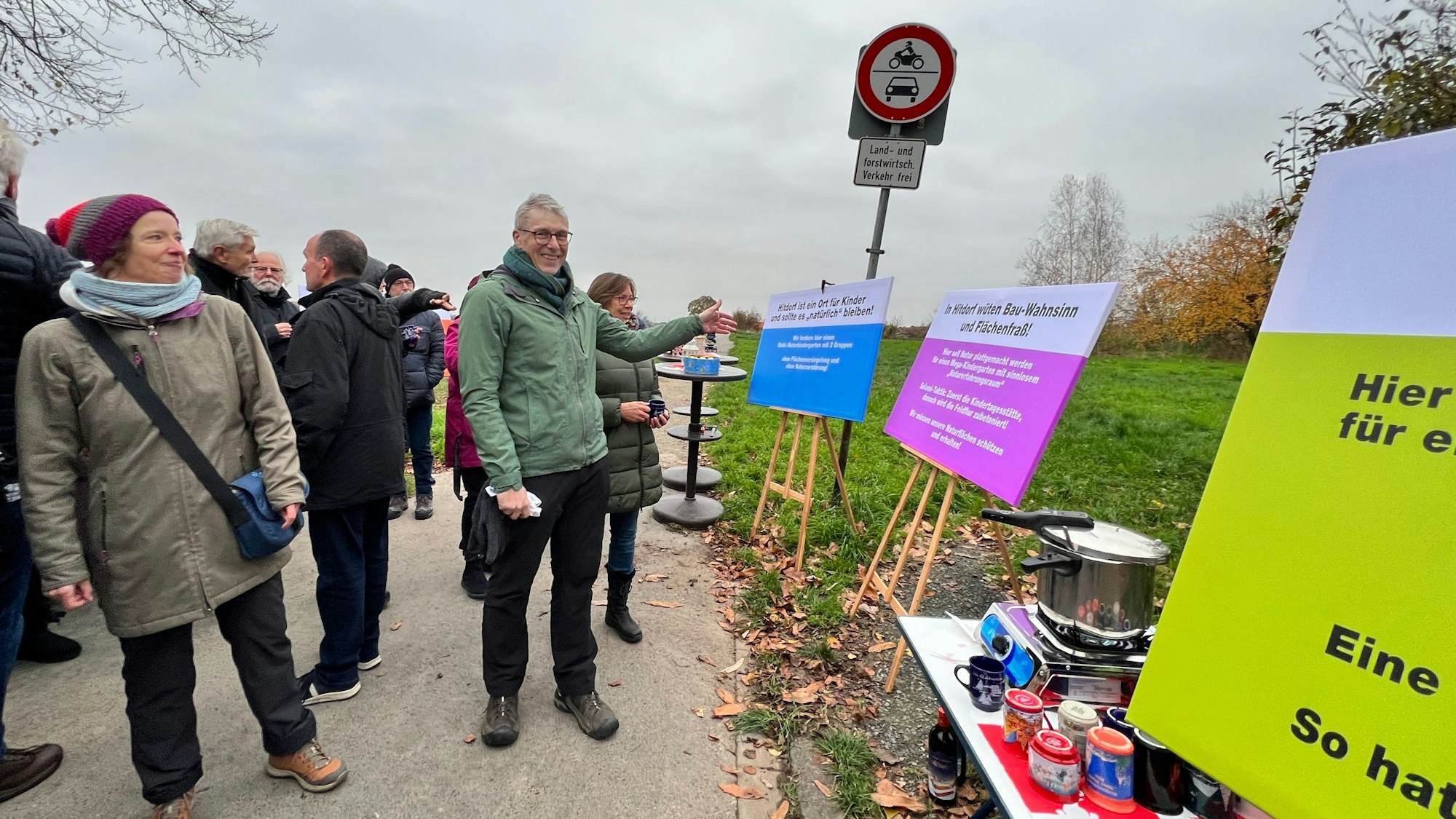 Links ist eine Gruppe von Menschen zu sehen. Einer von ihnen, Jörg Lichter, blickt in die Kamera und zeigt auf Plakat, die auf der rechten Seiten aufgetellt sind. Dort steht unter anderem „In Hitdorf wüten Bau-Wahnsinn und Flächenfraß!“