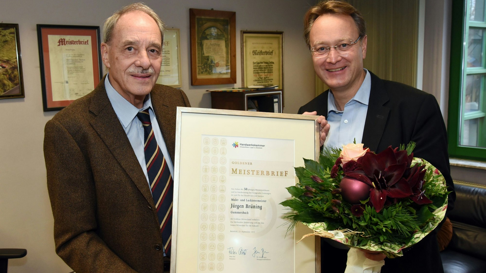 Zwei Männer im Sakko halten einen Rahmen mit einem Goldenen Meisterbrief in den Händen, einer hat in seiner anderen Hand einen Blumenstrauß.
