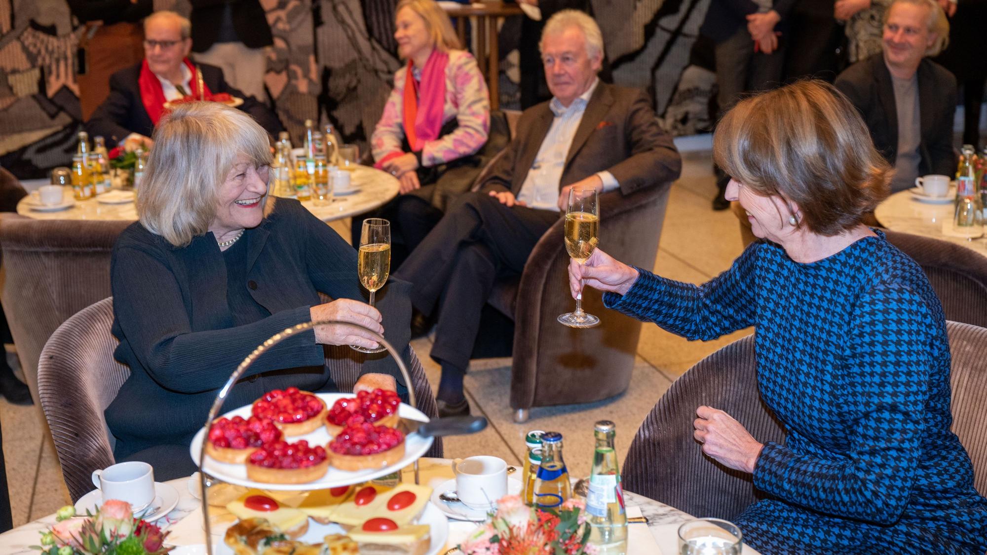 Alice Schwarzer und Henriette Reker am Tisch mit einem Glas Sekt in der Hand.