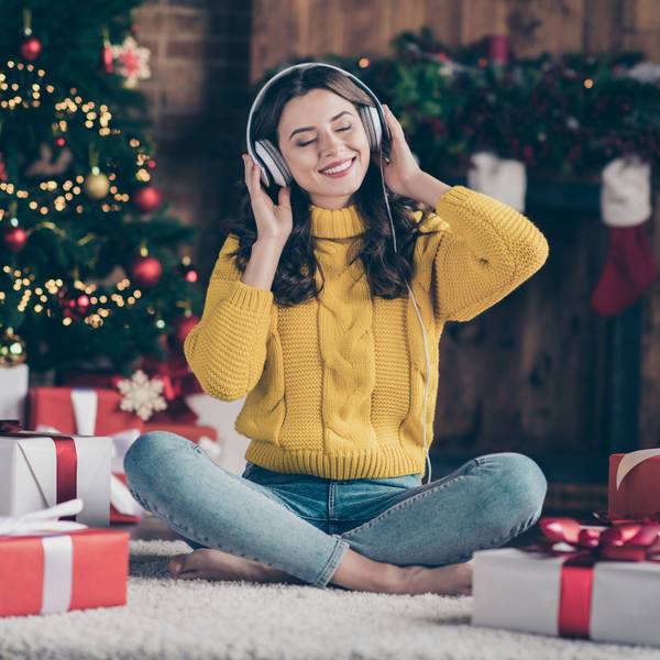 Eine Frau mit Kopfhörern sitzt vor dem Weihnachtsbaum und hat Geschenke vor sich stehen. Sie hört Musik und lächelt mit geschlossenen Augen.