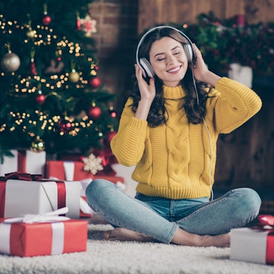 Eine Frau mit Kopfhörern sitzt vor dem Weihnachtsbaum und hat Geschenke vor sich stehen. Sie hört Musik und lächelt mit geschlossenen Augen.