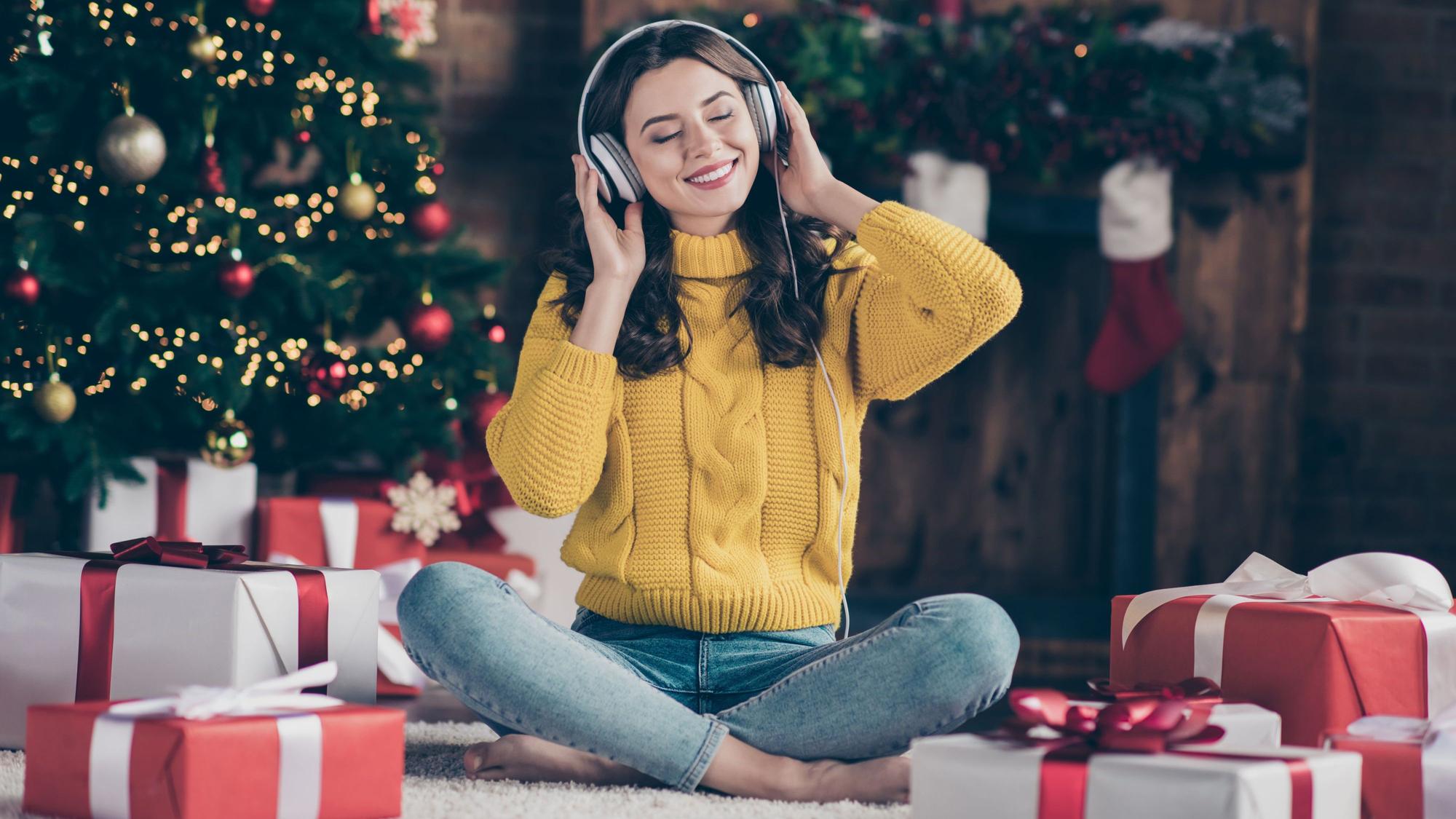 Eine Frau mit Kopfhörern sitzt vor dem Weihnachtsbaum und hat Geschenke vor sich stehen. Sie hört Musik und lächelt mit geschlossenen Augen.
