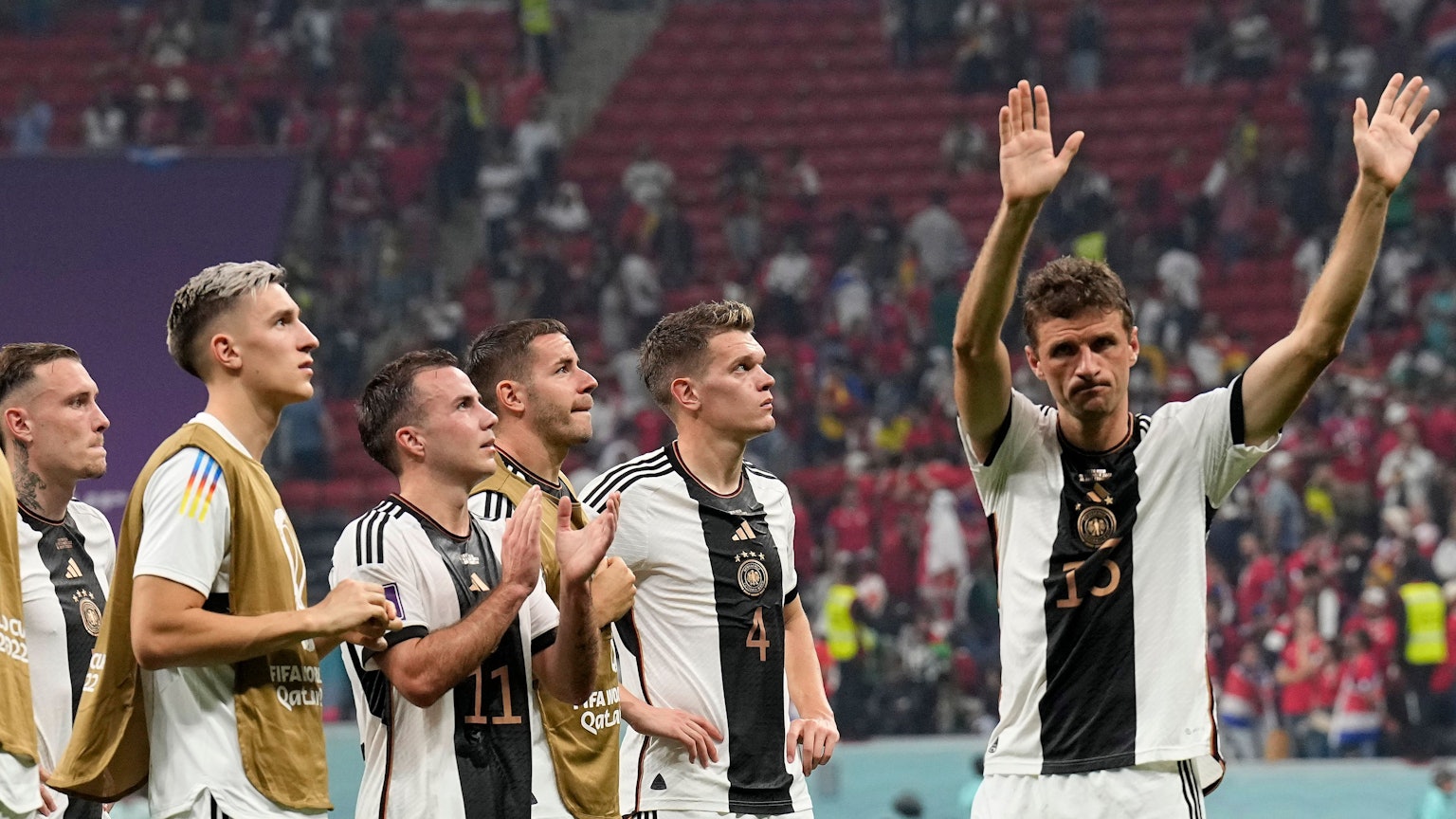 Im Stadion in Katar stehen sechs Spieler der deutschen Nationalmannschaft enttäuscht auf dem Spielfeld vor bereits halbleeren Rängen. Fünf von ihnen schauen nach oben, vermutlich auf die Anzeigentafel, die den Punktestand der Vorrundengruppe zeigt. Rechts winkt Thomas Müller Fans zum Abschied zu.