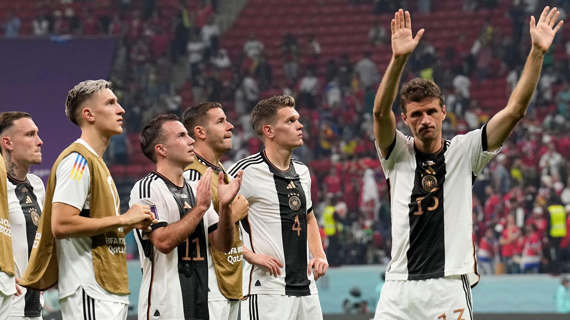 Im Stadion in Katar stehen sechs Spieler der deutschen Nationalmannschaft enttäuscht auf dem Spielfeld vor bereits halbleeren Rängen. Fünf von ihnen schauen nach oben, vermutlich auf die Anzeigentafel, die den Punktestand der Vorrundengruppe zeigt. Rechts winkt Thomas Müller Fans zum Abschied zu.