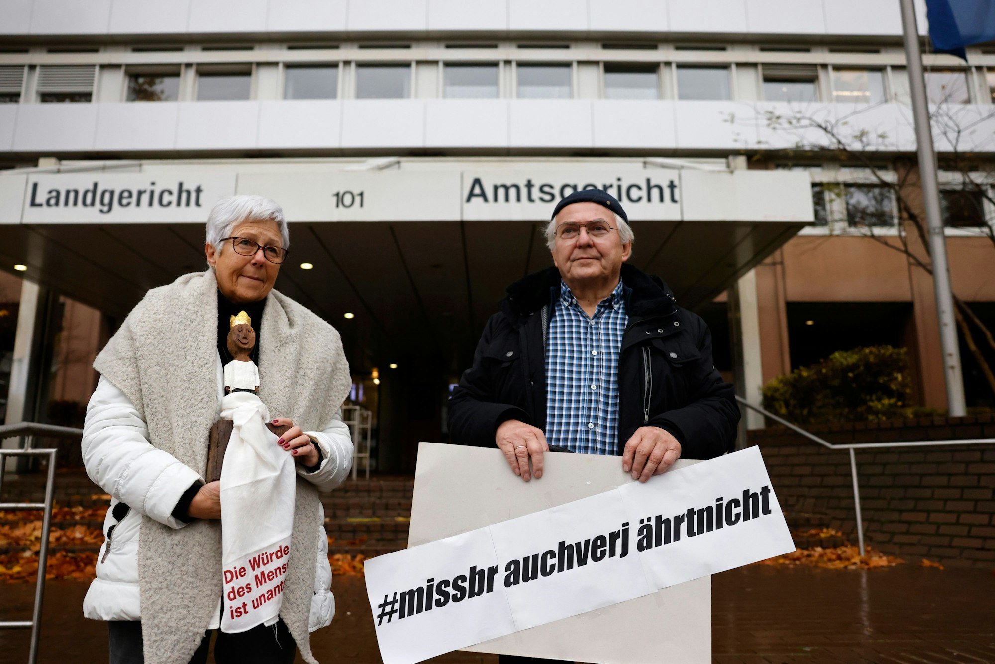 Eine Frau und ein Mann stehen vor dem Gericht mit dem Plakat in der Hand.