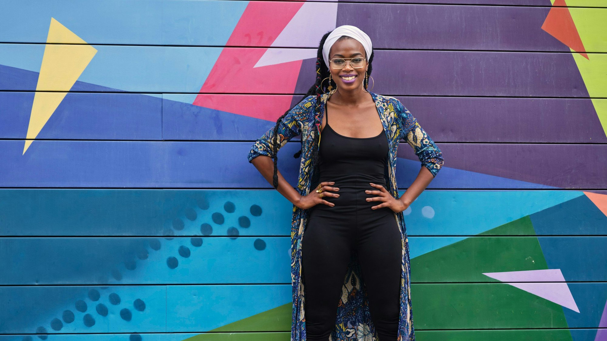 Sängerin Melane Nkounkolo vor einem Afroshop, Köln. Sie hat die Hände auf die Hüften gestemmt, der Hintergrund ist mit bunten geometrischen Mustern bemalt.