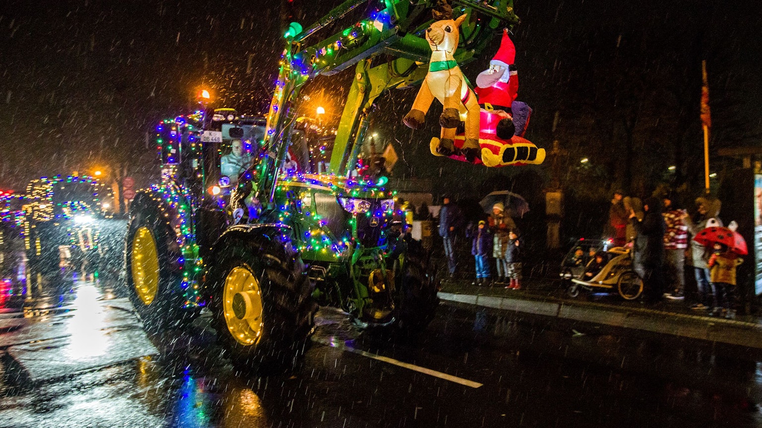 Ein mit grünen und lila-farbenen Lichtern geschmückter Trecker mit einer beleuchteten Weihnachtsmann-Figur und einem Rentier fährt des Nachts auf einer Straße, am Rand stehen Zuschauer.