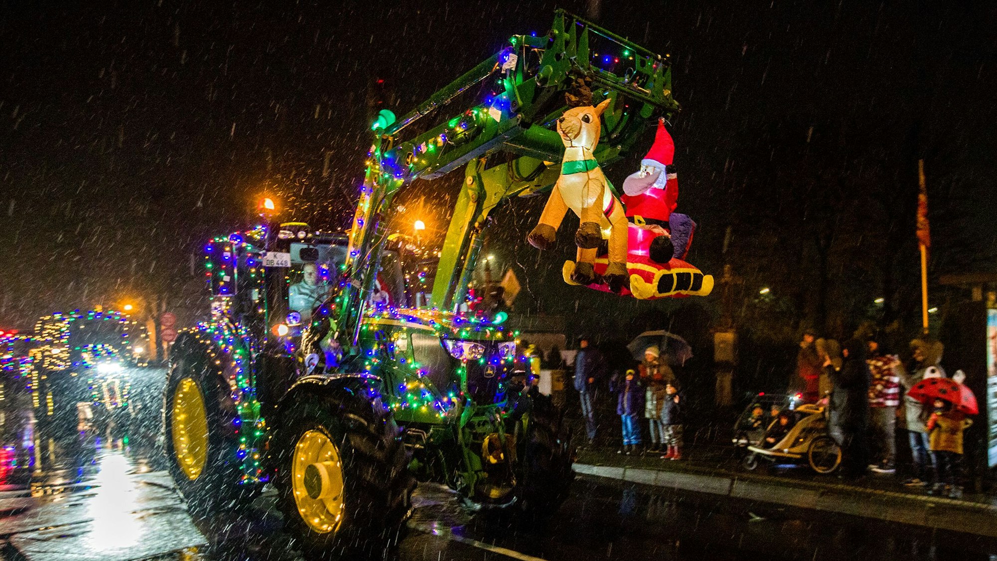 Ein mit grünen und lila-farbenen Lichtern geschmückter Trecker mit einer beleuchteten Weihnachtsmann-Figur und einem Rentier fährt des Nachts auf einer Straße, am Rand stehen Zuschauer.