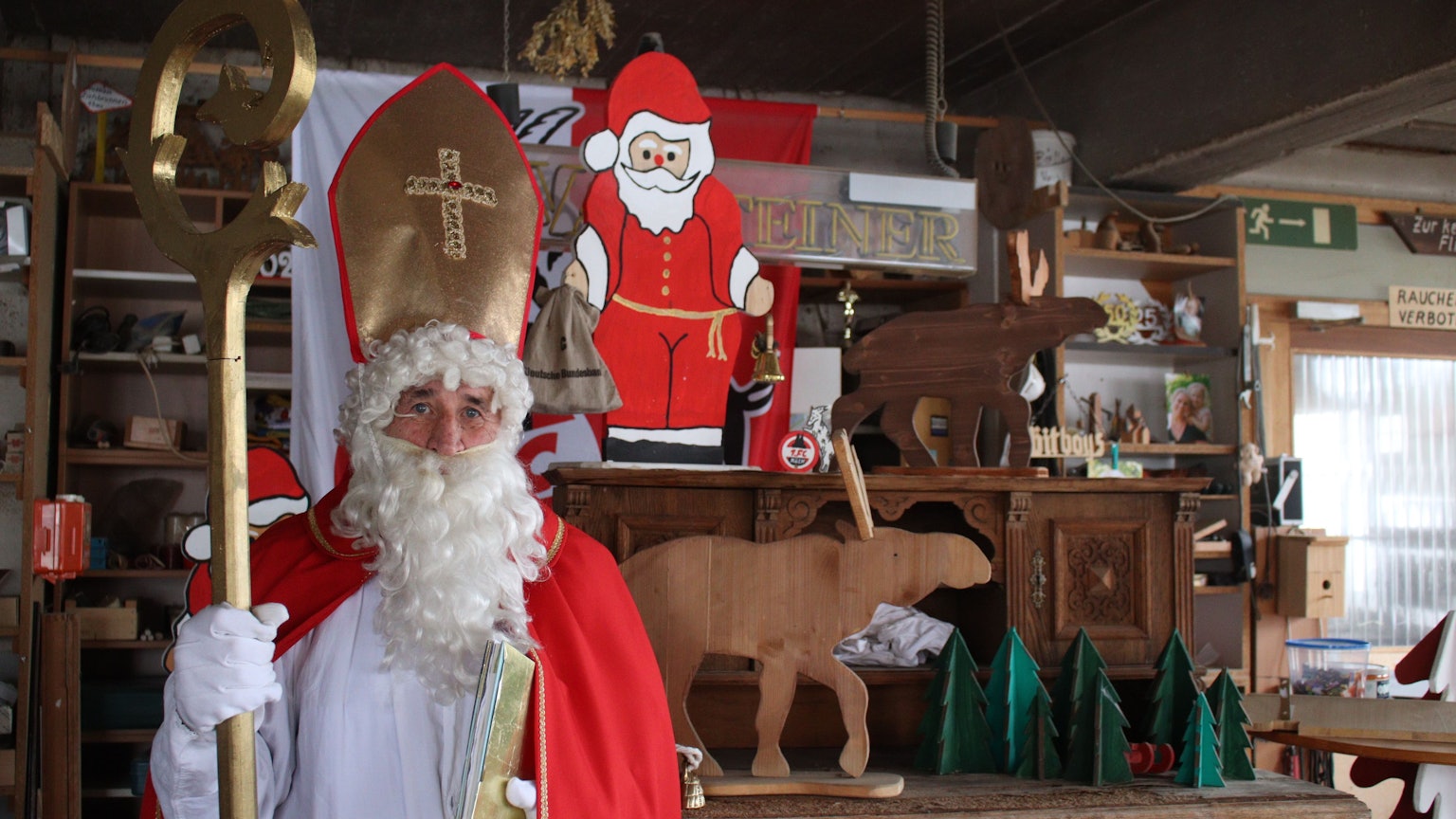 Hans-Peter Lückerath, genannt Hacky, steht im Nikolausgewand mit Stab und goldenem Buch in der Hand in seiner Schreinerwerkstatt in Nöthen. Im Hintergrund befinden sich unter anderem ein Nikolaus, Elche und Weihnachtsbäume aus Holz.