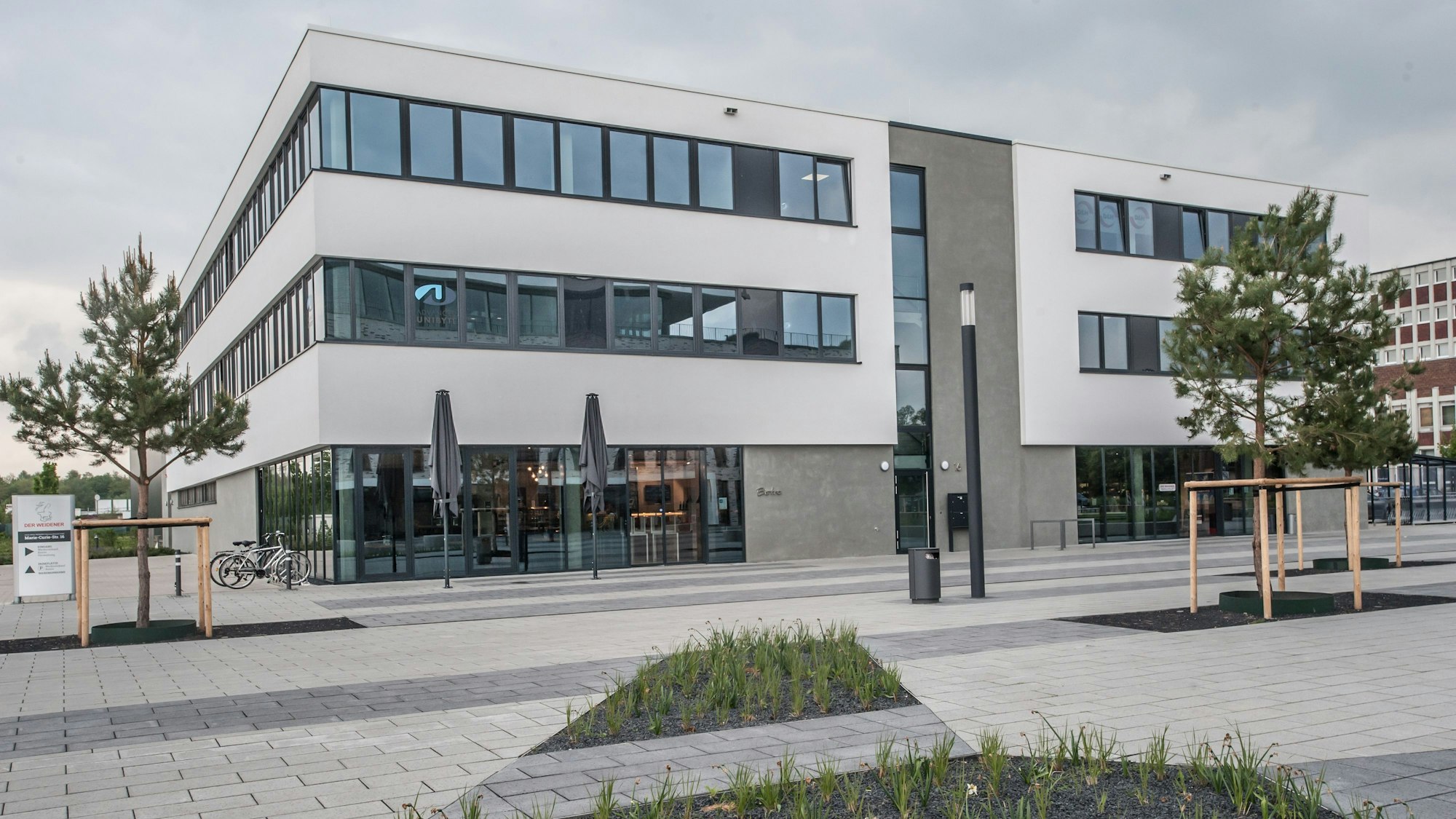 Ein gepflegtes Beet, eine neue gepflasterter Fläche, dahinter erhebt sich ein modernes Bürogebäude. Im Erdgeschoss ist ein Fleischhandel untergebracht.