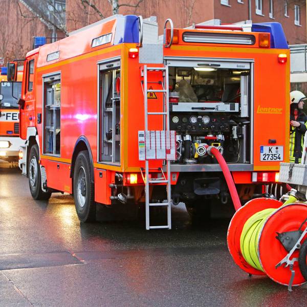 Die Kölner Feuerwehr rückt bei Brandstiftung aus (Symbolbild). Zwei Einsatzfahrzeuge stehen auf der Straße mit zwei Feuerwehrmännern.