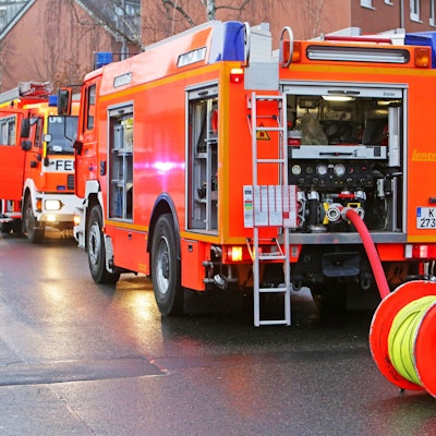 Die Kölner Feuerwehr rückt bei Brandstiftung aus (Symbolbild). Zwei Einsatzfahrzeuge stehen auf der Straße mit zwei Feuerwehrmännern.