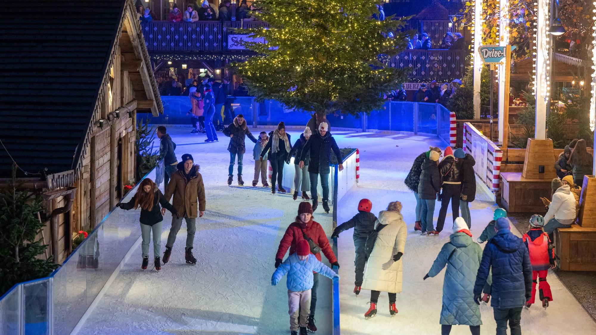 Eisbahn mit Schlittschuhläufern auf zwei Fahrbahnen, links eine Holhütte, in der Mitte ein geschmückter Tannenbaum