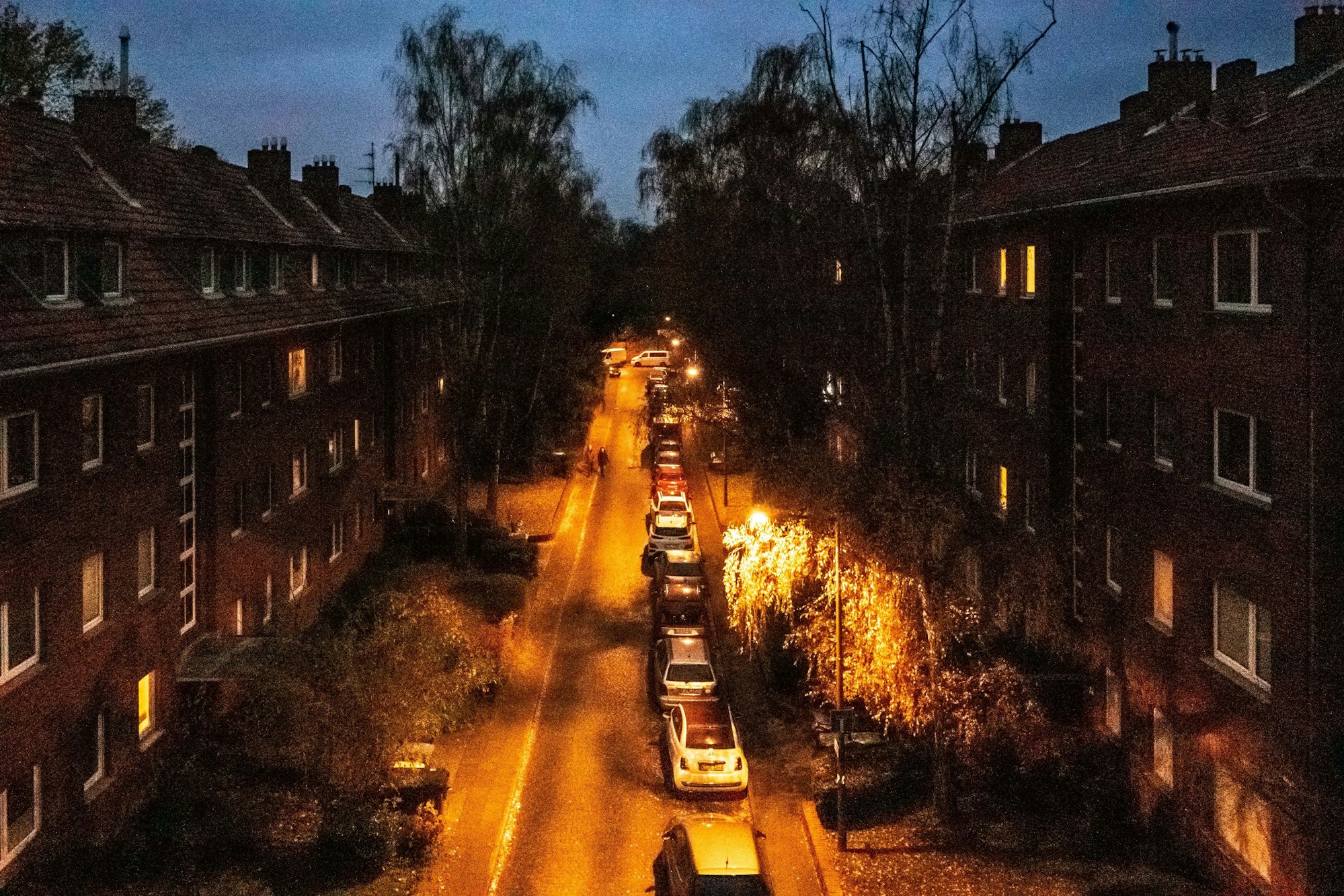 Eine Straße mit geparkten Autos und zwei Häuserreihen ist zu sehen, Straßenlaternen erleuchten sie.