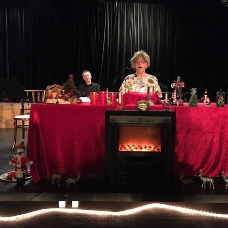 Ein Mann mit Brille sitzt hinter einem altarartigen Tisch mit roter Samtdecke, der weihnachtlich dekoriert ist, neben ihm ist ein Weihnachtsbaum aus Holz zu sehen, im Hintergrund sitzt ein weiterer Mann.