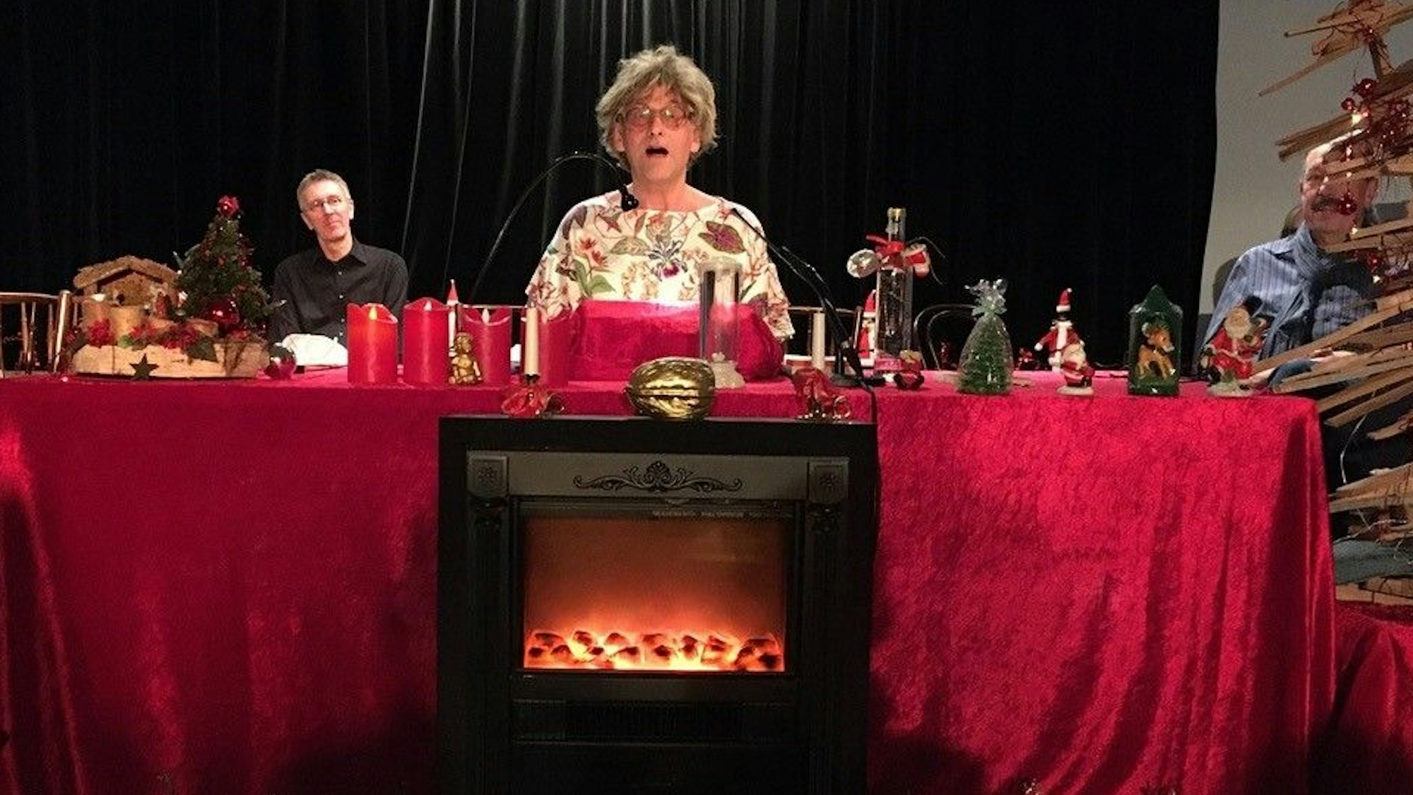 Ein Mann mit Brille sitzt hinter einem altarartigen Tisch mit roter Samtdecke, der weihnachtlich dekoriert ist, neben ihm ist ein Weihnachtsbaum aus Holz zu sehen, im Hintergrund sitzt ein weiterer Mann.