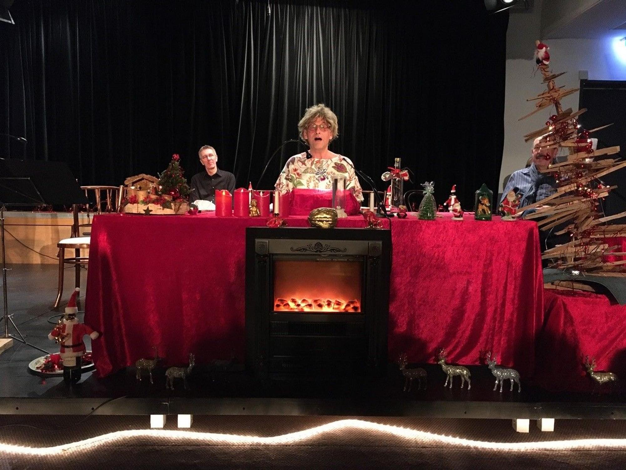 Ein Schauspieler sitzt auf einer weihnachtlich geschmückten Bühne und trägt etwas vor.